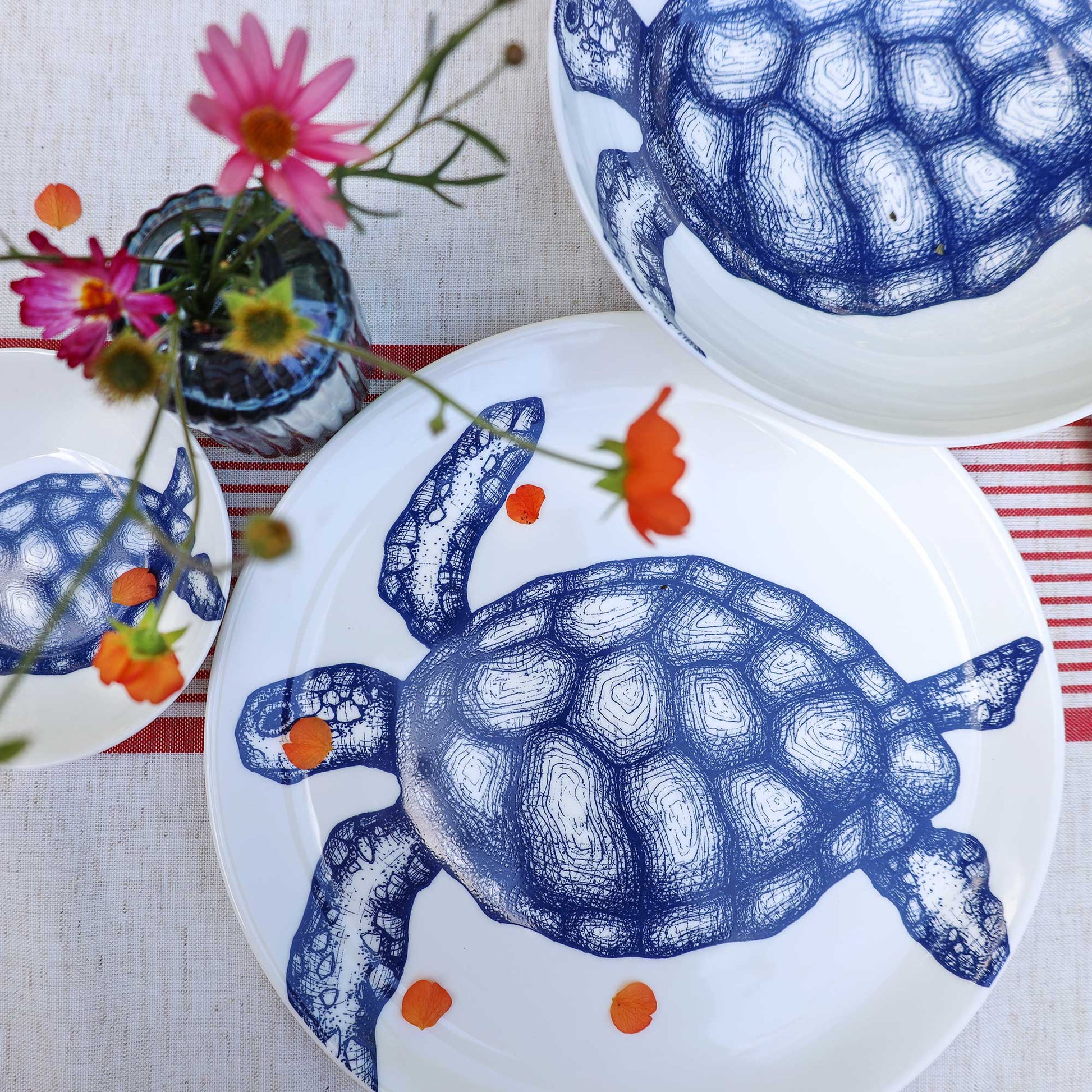 White ceramic plates decorated with a blue turtle illustration, arranged on a striped table runner with flowers in a glass vase.