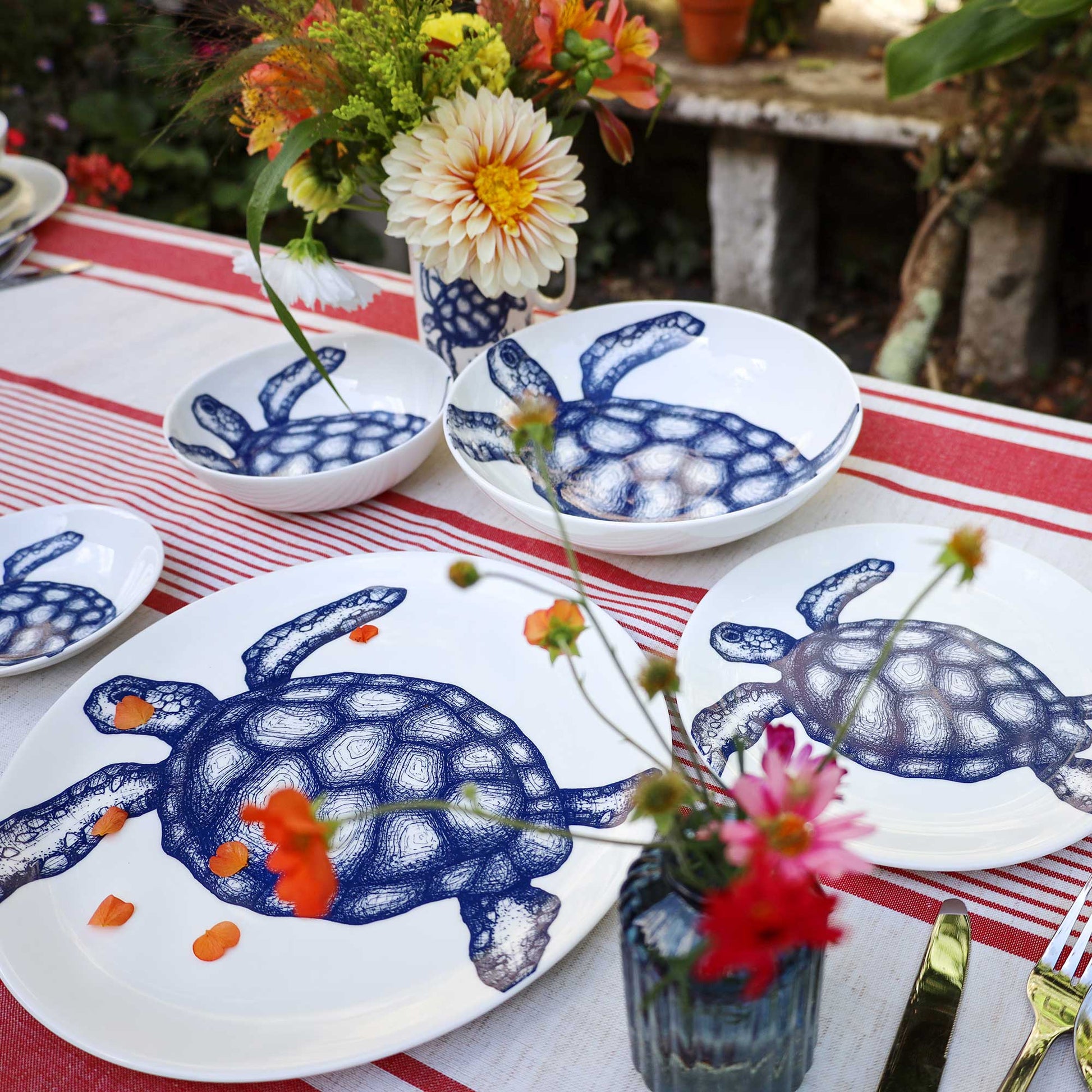 White ceramic tableware set featuring blue turtle illustrations, arranged on a red-and-white striped tablecloth with flowers in vases in an outdoor garden setting.
