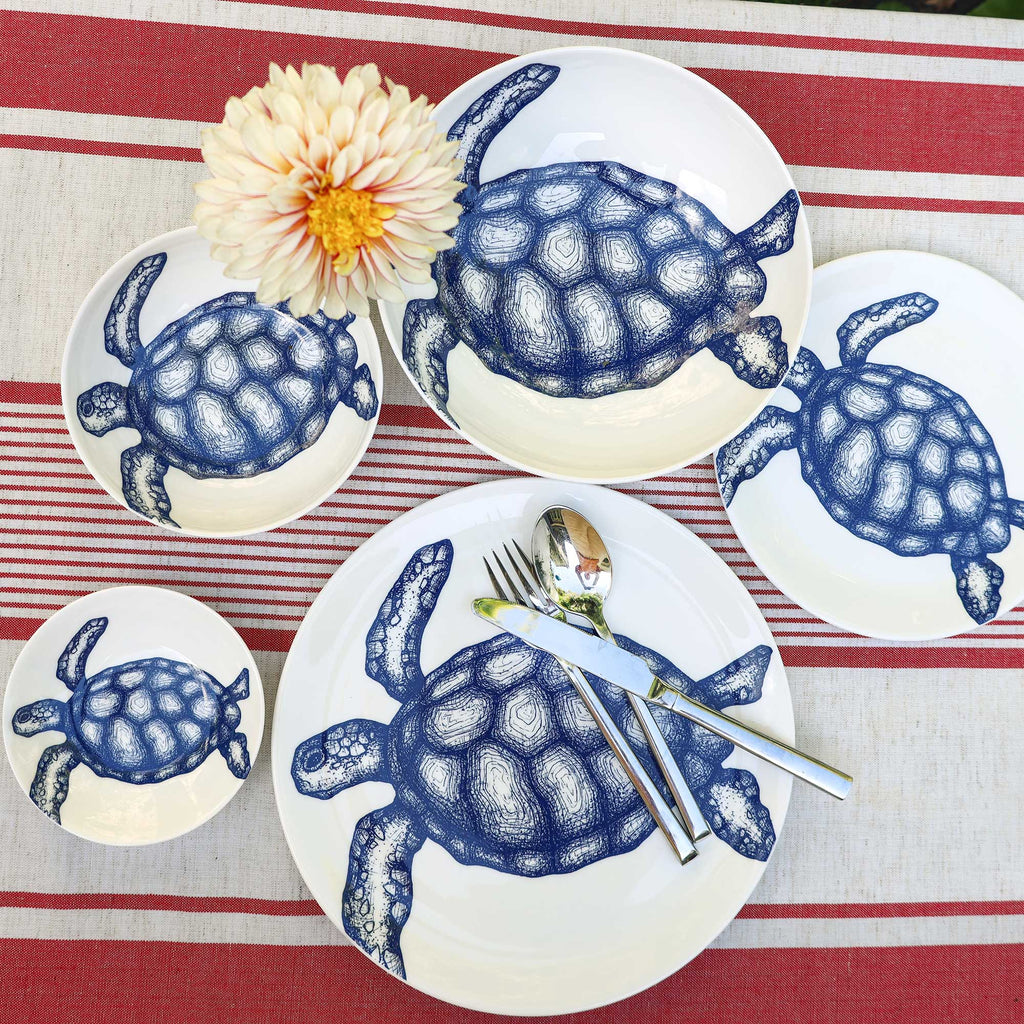 A set of white ceramic plates and bowls decorated with blue sea turtle illustrations, arranged on a red and white striped tablecloth with a white flower and silver cutlery.