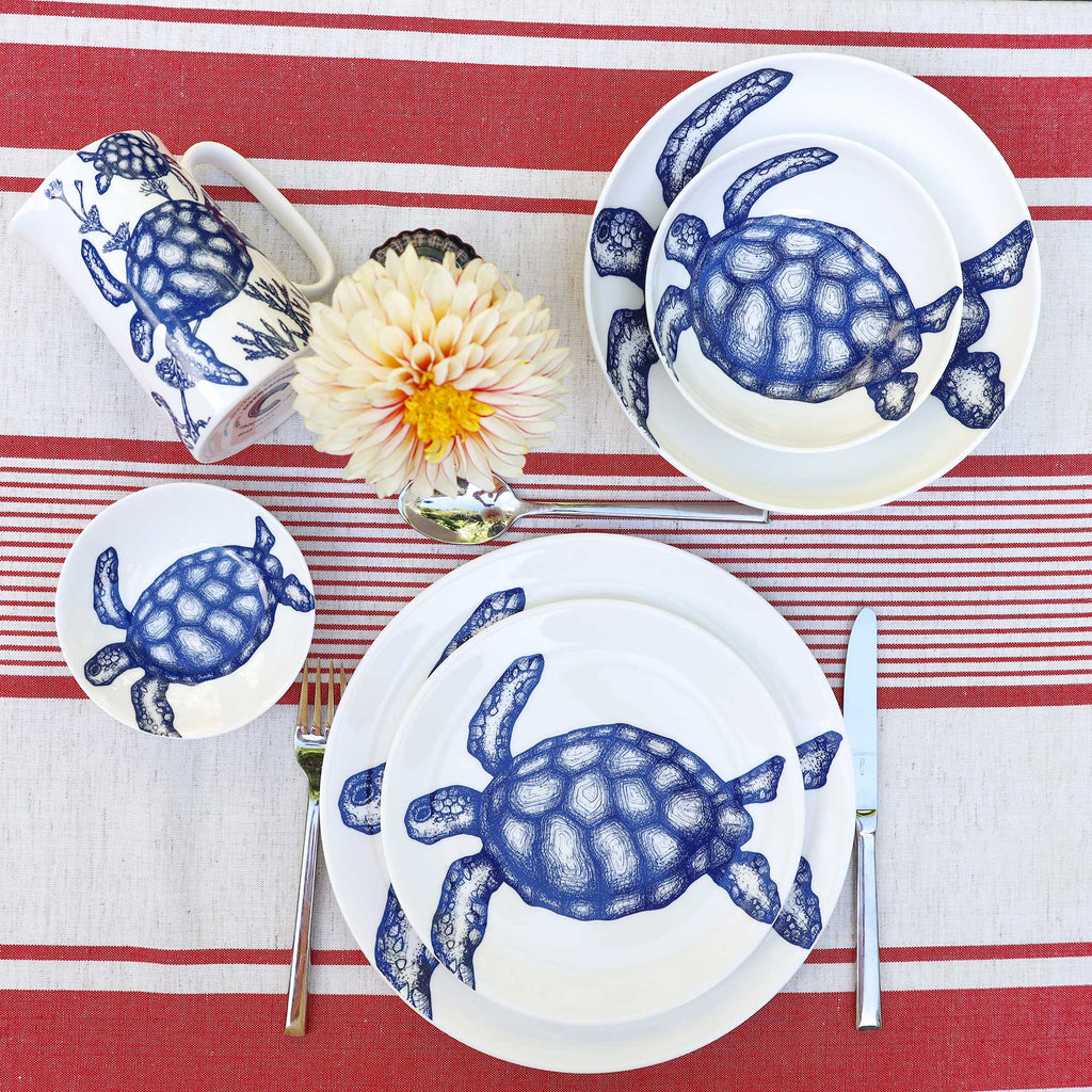A place setting featuring a white ceramic dinnerware set with a blue turtle print, arranged on a red and white striped tablecloth with a flower and silver cutlery.