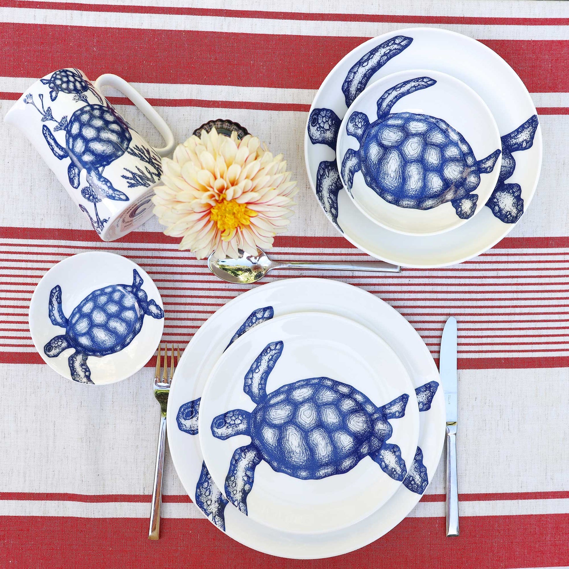 A place setting featuring a white ceramic dinnerware set with a blue turtle print, arranged on a red and white striped tablecloth with a flower and silver cutlery.