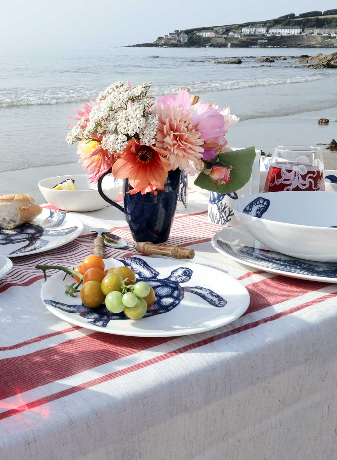 A styled beach picnic setup on a white and red striped tablecloth with blue-and-white ceramic dishes, a wicker picnic basket, flowers, bread, and red tomatoes, set against a sandy shoreline and ocean water.