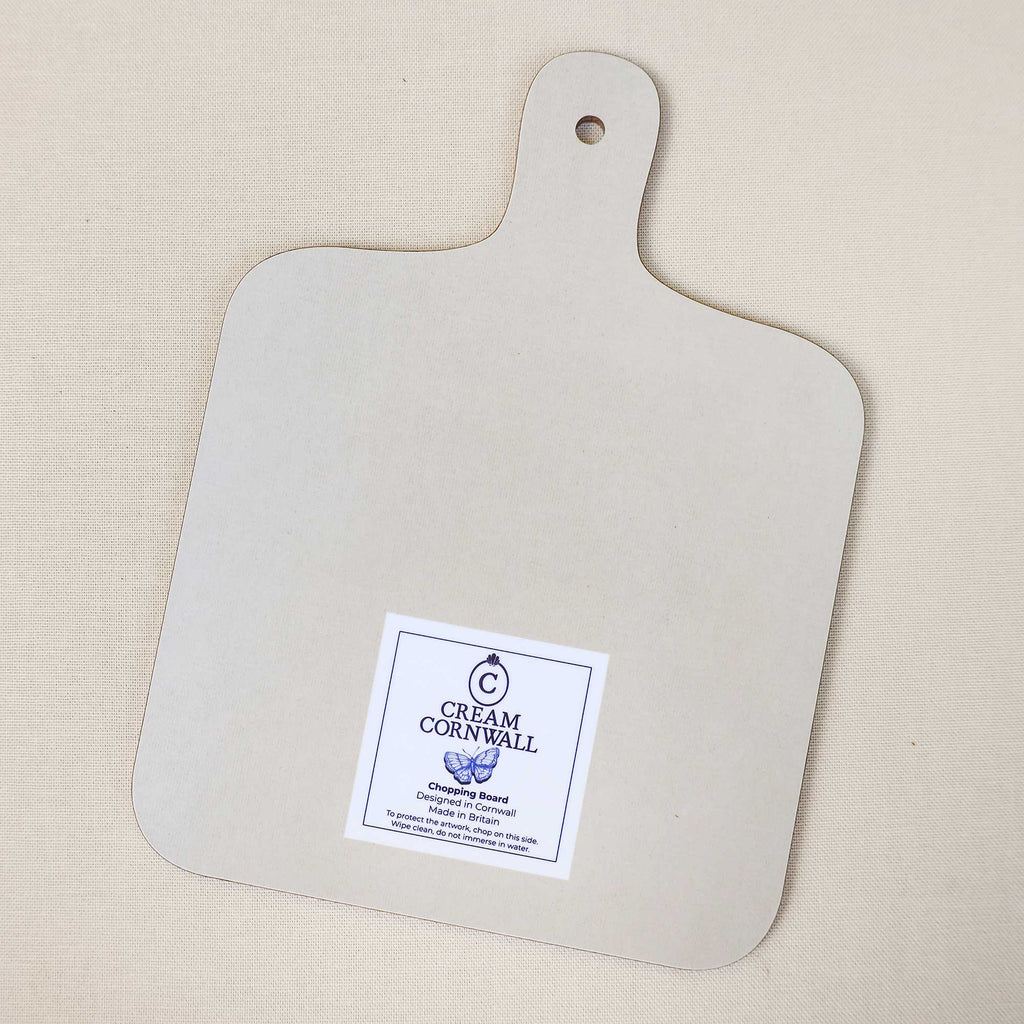 Reverse side of a Cream Cornwall chopping board, featuring a plain cream surface, rounded corners and a handle with a hanging hole. A Cream Cornwall branded care label is positioned near the bottom, photographed on a neutral linen background.