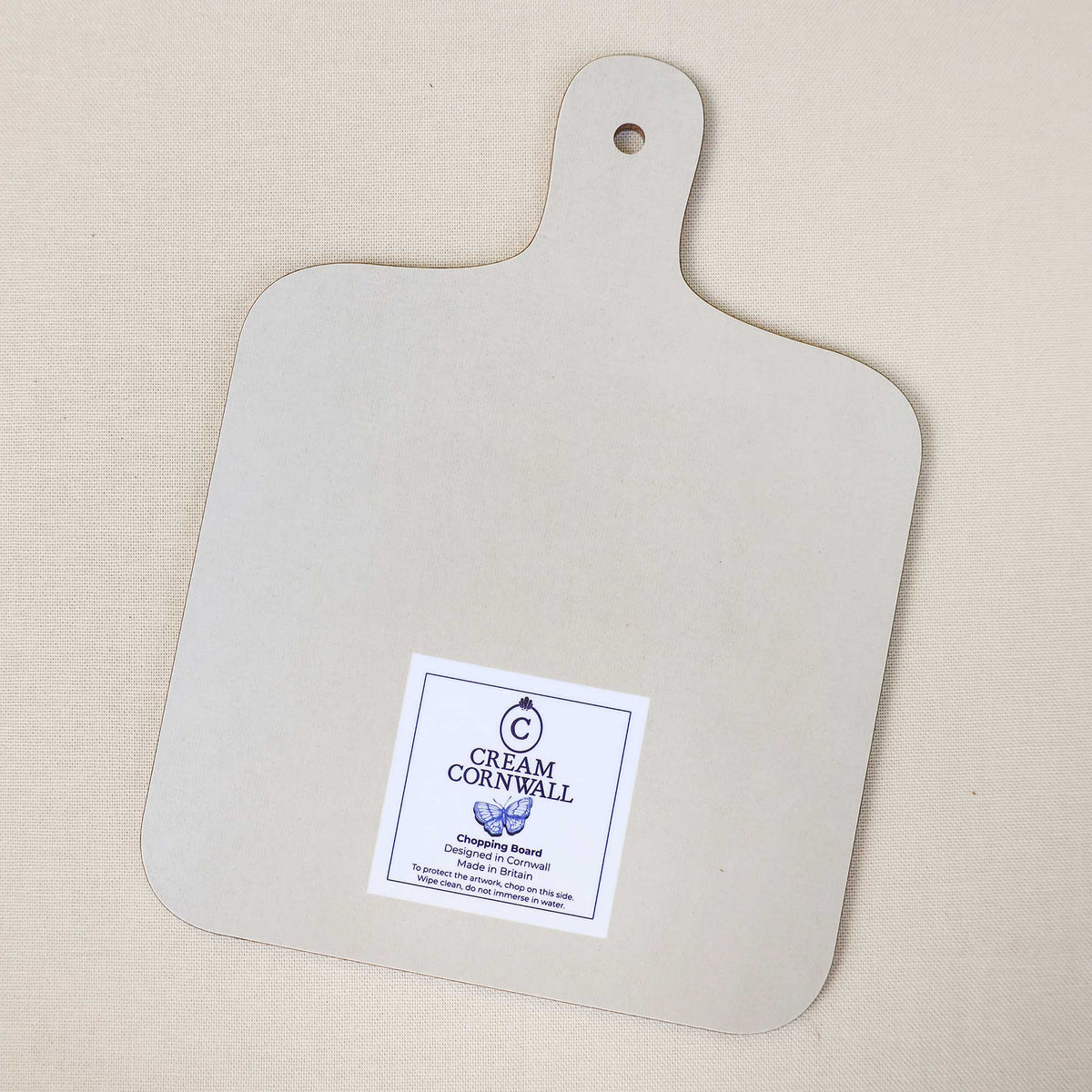 Reverse side of a Cream Cornwall chopping board, featuring a plain cream surface, rounded corners and a handle with a hanging hole. A Cream Cornwall branded care label is positioned near the bottom, photographed on a neutral linen background.
