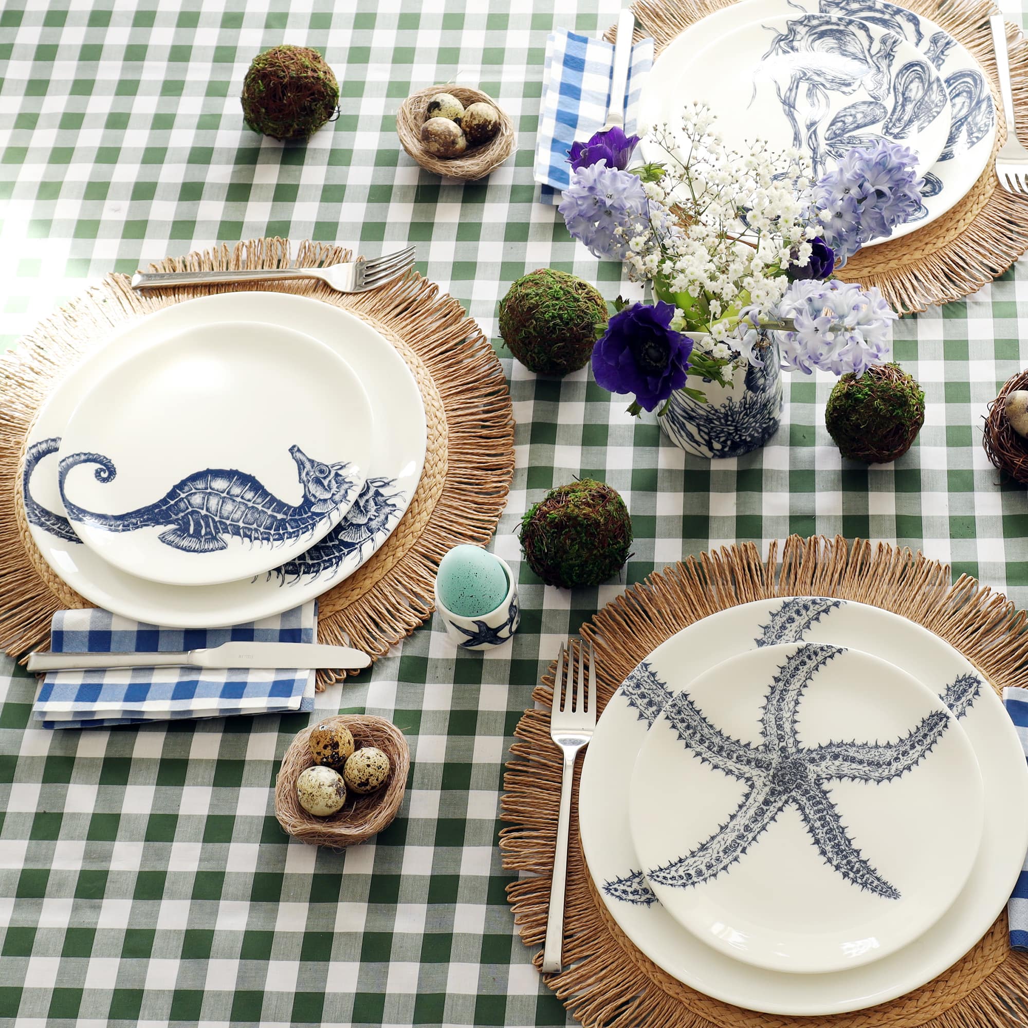 White bone china dinner plate with side plate sitting on top. Bothe plate have a navy blue illustration of a starfish on. They are set as a place setting on a raffia placemat with blue gingham napkin and green gingham tablecloth. the table is set for Easter with eggs in egg cups and moss balls.