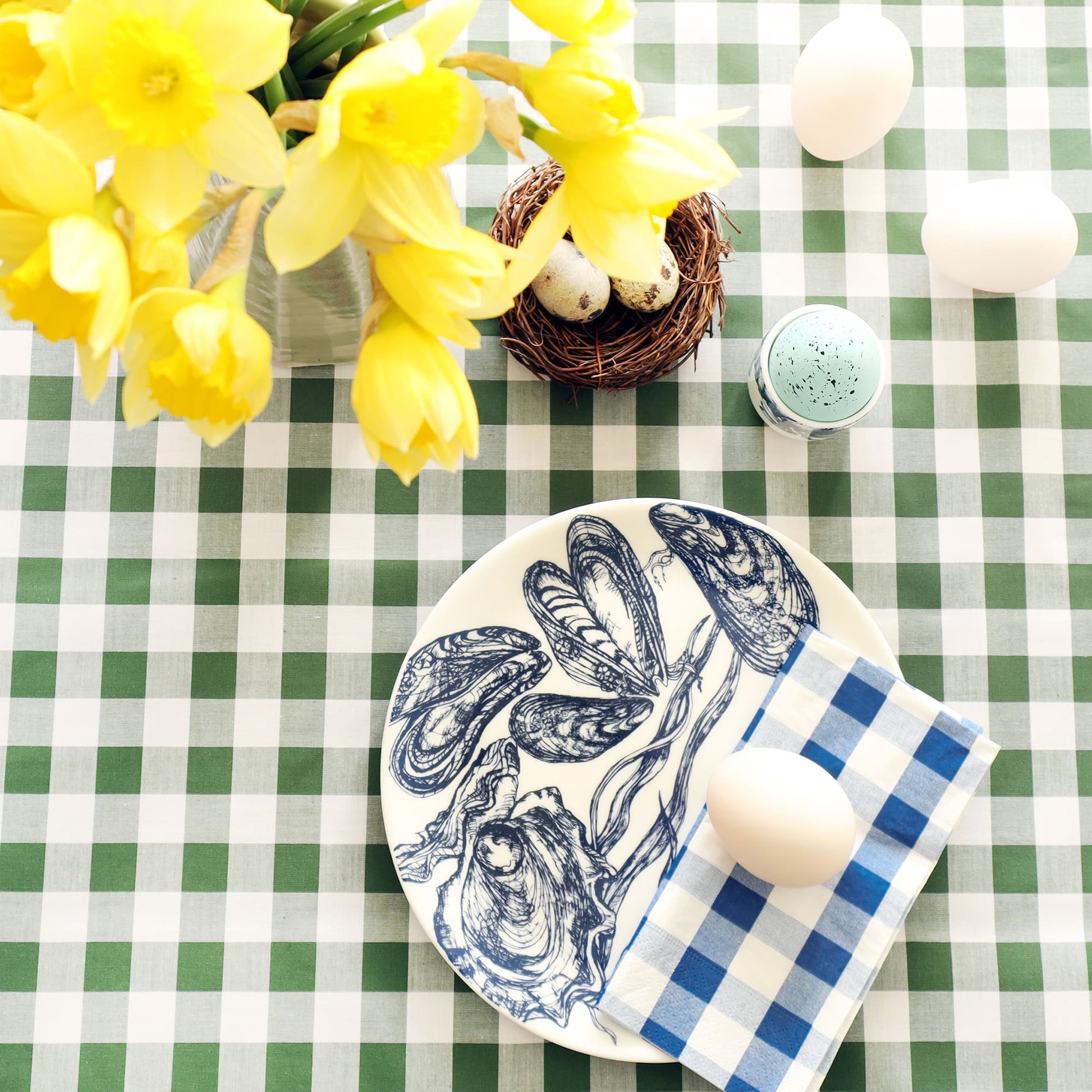 White plate with navy blue mussels & oysters decoration with folded blue gingham napkin and duck egg on it. This is on a green gingham tablecloth with a small nest and quails eggs, hyacinths and anemones and more eggs.