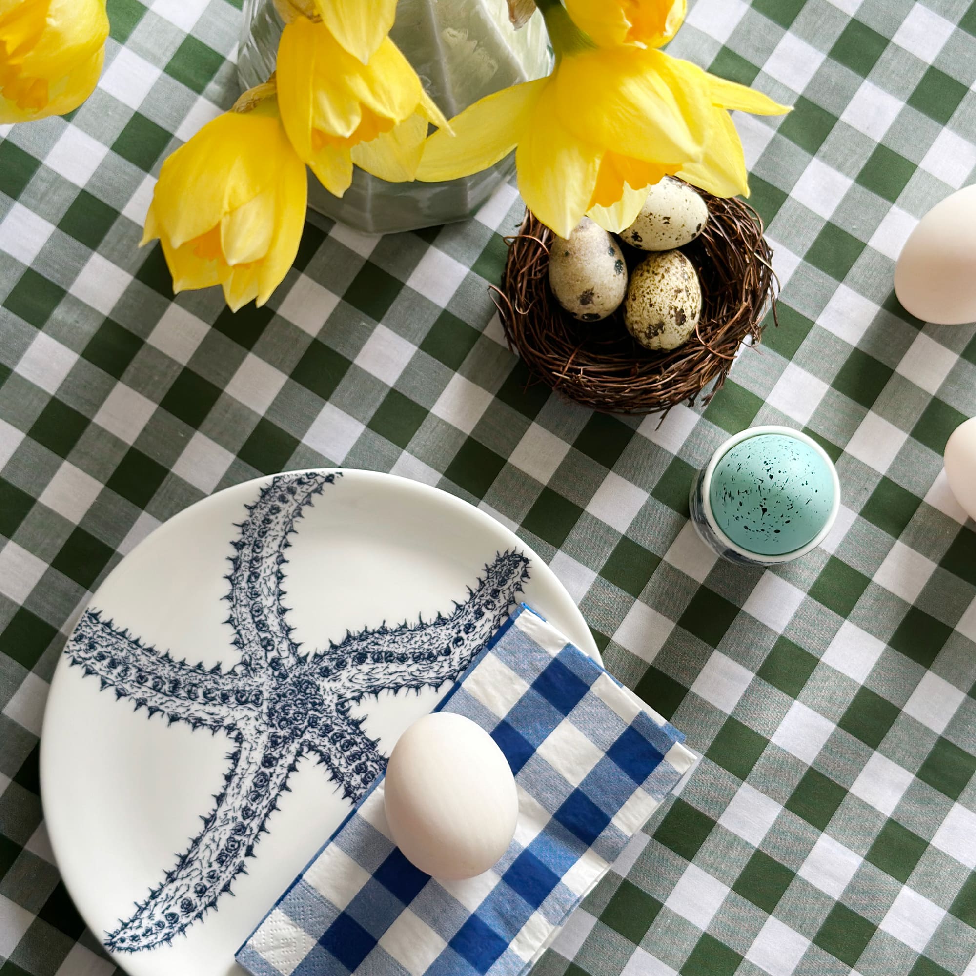 White bone china pate with navy illustrated starfish on. The plate is on a green gingham tablecloth with eggs and daffodils set for Easter.
