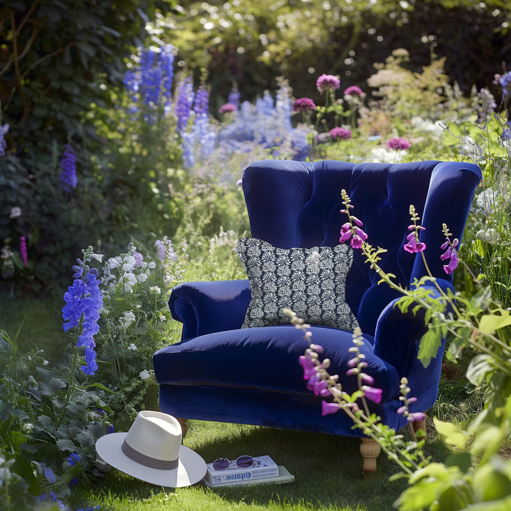 A deep blue velvet armchair sits in the middle of a vibrant garden, surrounded by blooming purple, pink, and blue flowers. The chair is styled with a navy cushion featuring a white coral-like pattern. On the grass nearby rests a white sunhat, a book, and a pair of sunglasses, evoking a peaceful, summery reading nook.