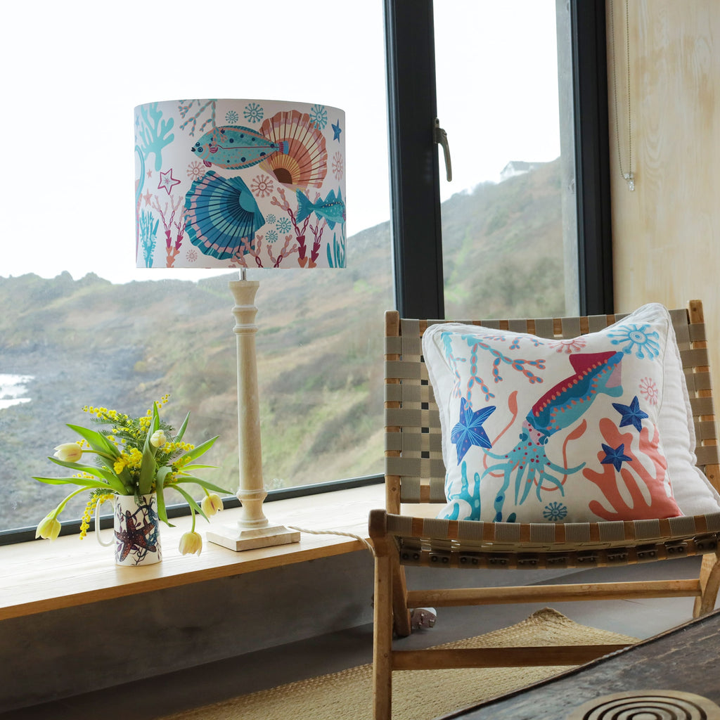 A cozy coastal-inspired interior with a wooden chair featuring a marine-themed cushion decorated with colorful illustrations of a flat fish, seashells, and coral in shades of blue, pink, and orange. Beside the chair, a wooden lamp with a matching ocean-inspired lampshade sits on a windowsill overlooking a rugged coastal landscape. A decorative mug filled with fresh yellow tulips adds a natural touch to the setting.