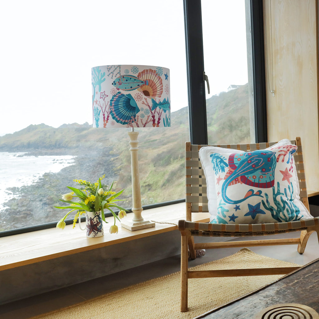 A bright and airy coastal interior featuring a wooden chair with a woven seat, adorned with a marine-themed cushion showcasing a vibrant stingray design in shades of blue, pink, and coral. Beside the chair, a wooden sideboard holds a tall lamp with a matching ocean-inspired lampshade decorated with fish, seashells, and coral. A white ceramic mug filled with fresh yellow tulips and greenery adds a natural touch. Large windows in the background offer a stunning view of a rocky coastline and rolling hills.