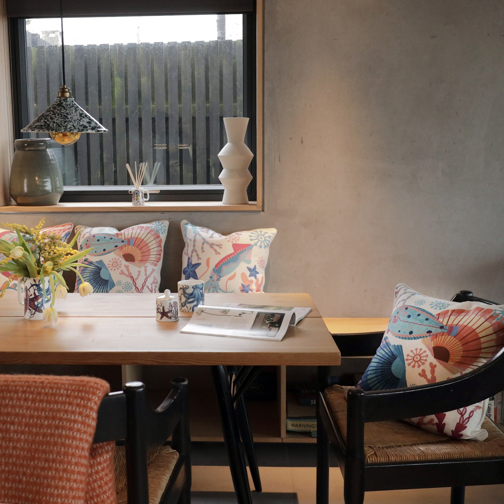 Cozy dining space with a wooden table, marine-themed cushions, and coastal decor. A vase of fresh flowers and an open magazine add a relaxed touch.