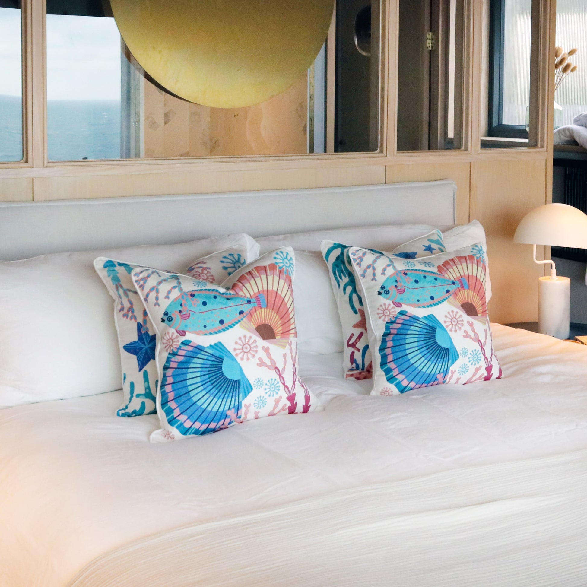 A serene coastal-themed bedroom featuring a neatly made bed with vibrant ocean-inspired cushions showcasing seashells, fish, and coral designs. The bed is dressed in crisp white linens, complemented by a sleek bedside lamp and a calming ambiance.
