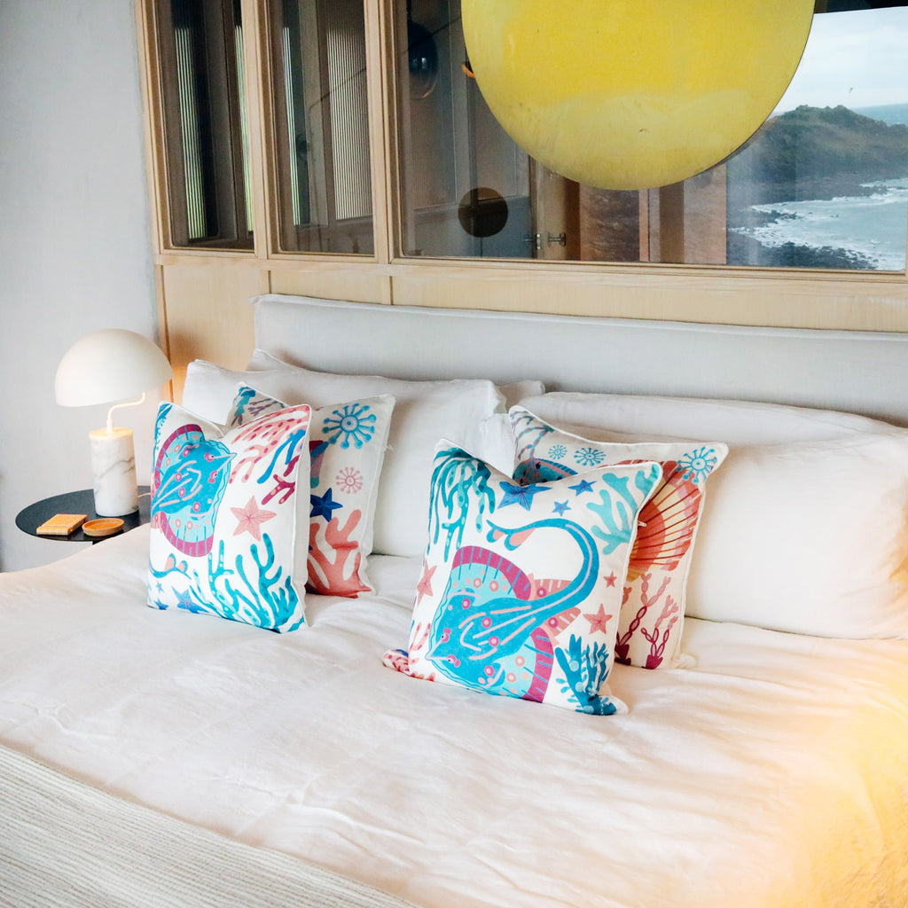 A cozy bedroom with a modern coastal theme, featuring a neatly made bed adorned with vibrant ocean-inspired cushions. The cushions showcase a bold marine design with stingrays, starfish, and coral in shades of blue, pink, and coral. The bed is dressed in crisp white linens, enhancing the fresh and airy ambiance. A sleek black bedside table holds a sculptural white lamp with a marble base. A large reflective yellow orb hangs above the bed, adding an artistic touch, while a window in the background offers a s