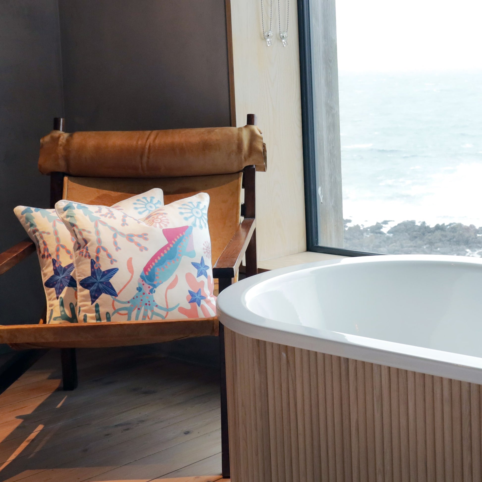 A serene bathroom setting with a freestanding bathtub near a large window overlooking a rocky coastline. A vintage-style leather armchair sits nearby, adorned with two marine-themed cushions featuring colorful illustrations of a squid, starfish, and coral in shades of blue, pink, and orange. The natural wood flooring and soft lighting add to the cozy, coastal ambiance.
