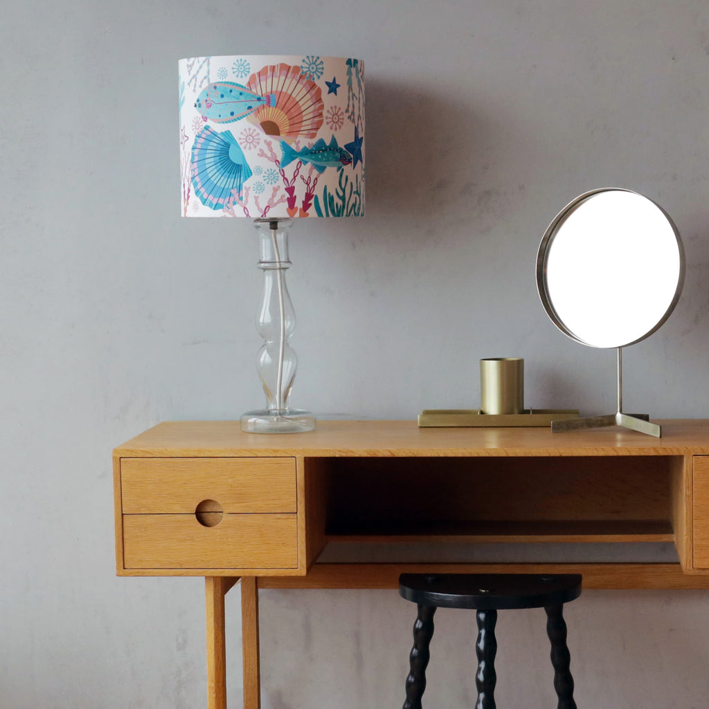 A minimalist wooden dressing table with a marine-themed lamp featuring a colorful lampshade adorned with illustrations of fish, seashells, and coral in shades of blue, pink, and orange. The lamp has a clear glass base. A round mirror with a metallic frame and a brass candle holder sit beside it, while a black stool with a twisted-leg design is tucked underneath the table. The setting is against a textured grey wall.