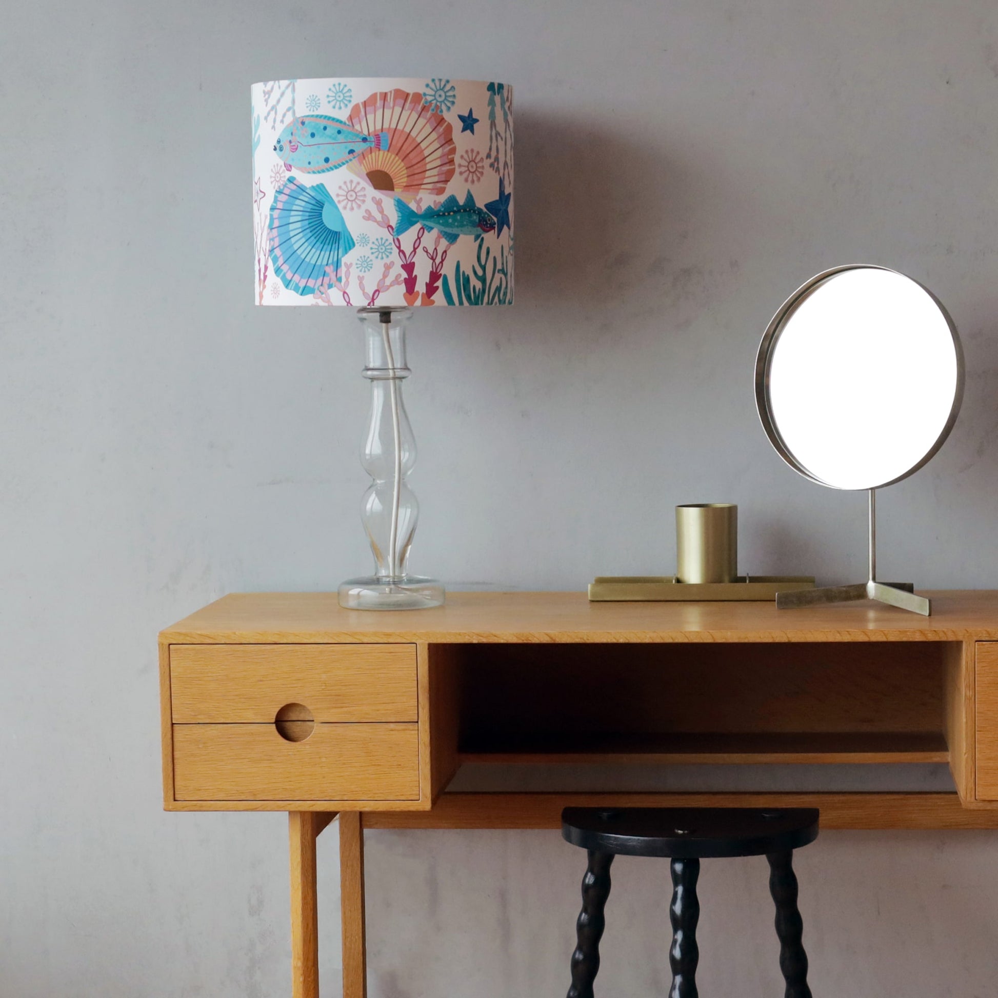 A minimalist wooden dressing table with a marine-themed lamp featuring a colorful lampshade adorned with illustrations of fish, seashells, and coral in shades of blue, pink, and orange. The lamp has a clear glass base. A round mirror with a metallic frame and a brass candle holder sit beside it, while a black stool with a twisted-leg design is tucked underneath the table. The setting is against a textured grey wall.