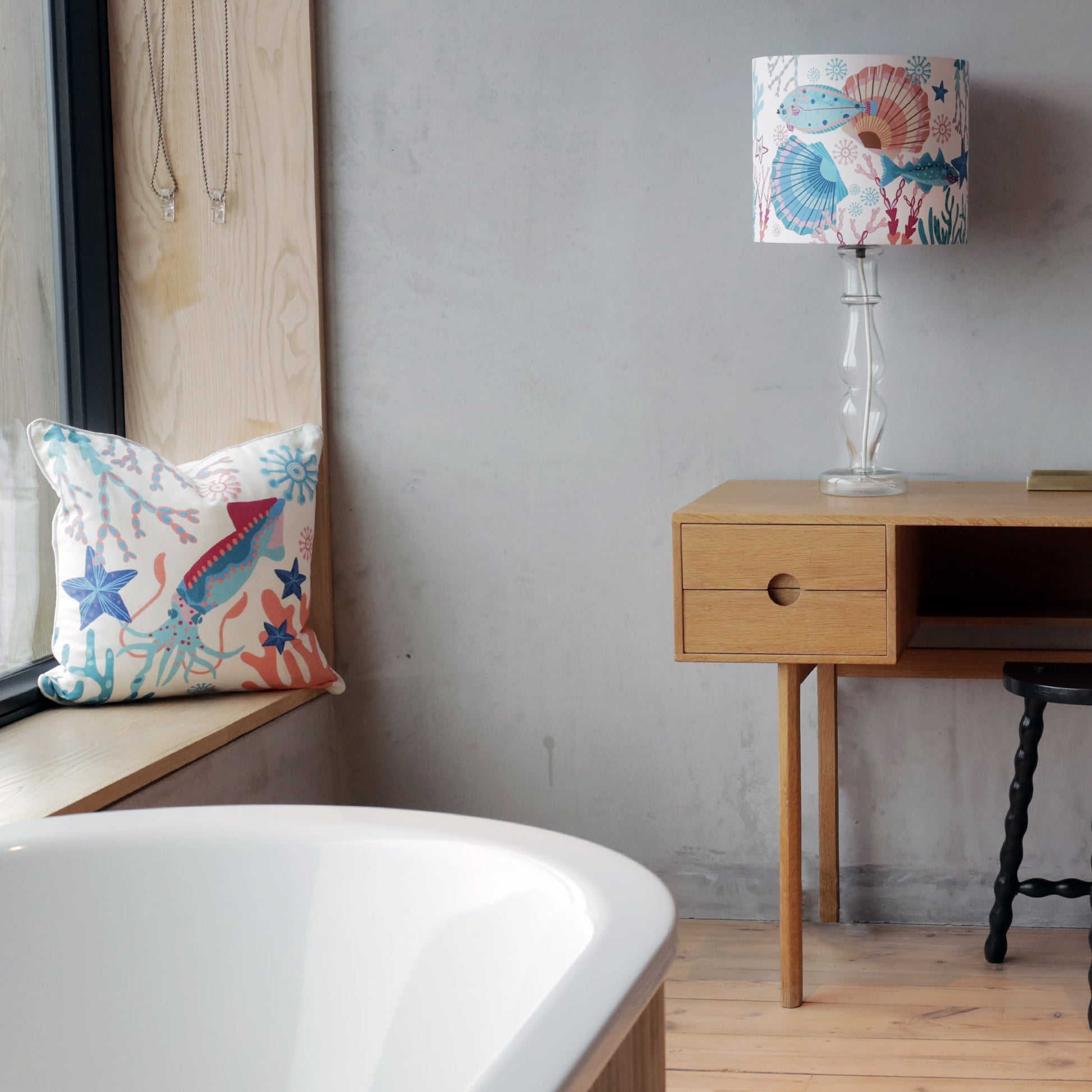 A cozy bedroom setting featuring a marine-themed cushion and matching lamp with colorful illustrations of a squid, fish, starfish, and coral in shades of blue, pink, and orange. The cushion rests on a white bed, while the lamp with a wooden base sits on a black bedside table. A brass candle holder and an open book complete the serene and coastal-inspired decor.