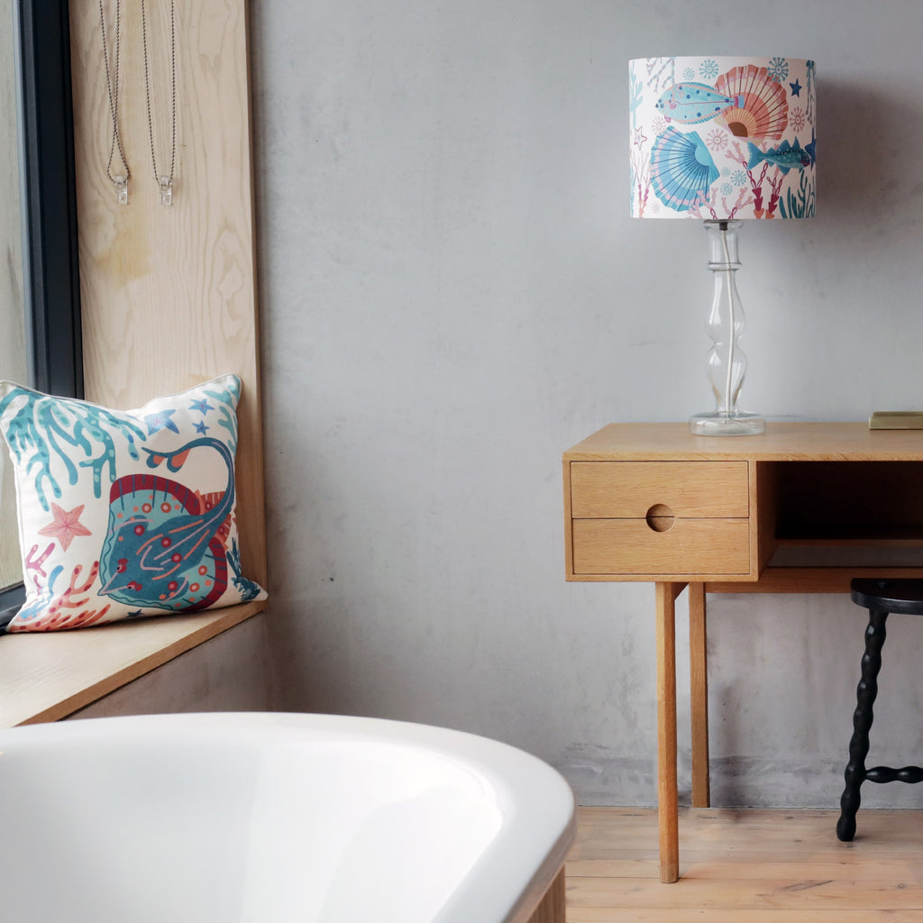 A serene bathroom setting featuring a modern freestanding bathtub in the foreground. A wooden bench by the window holds a vibrant ocean-themed cushion with an illustration of a stingray, coral, and starfish in shades of blue, red, and coral. A sleek wooden desk with a minimalist design stands against a textured concrete wall, accompanied by a black stool. On the desk, a tall glass lamp with a marine-inspired lampshade, adorned with seashell and fish illustrations, adds a coastal touch. 