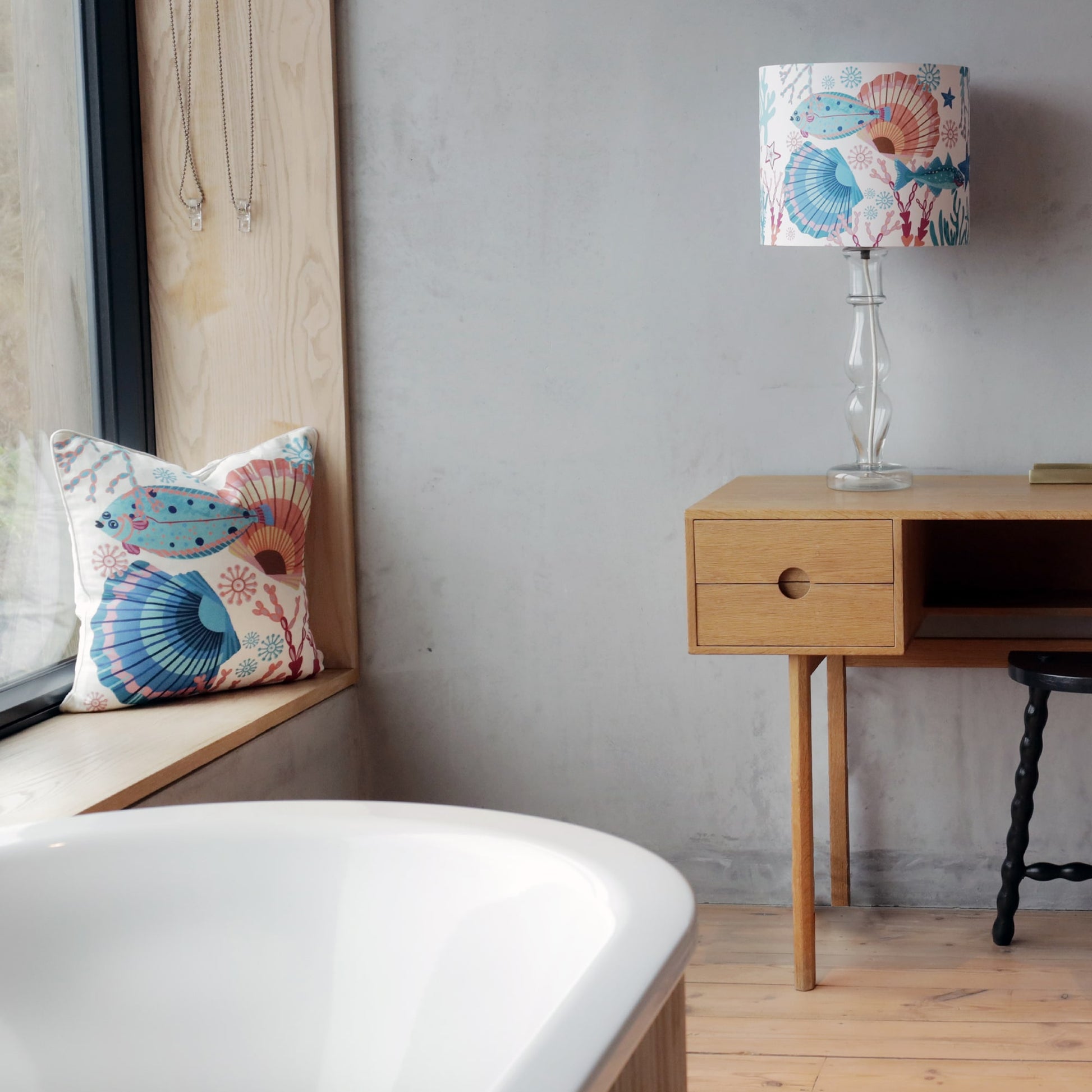 A minimalist bathroom with a freestanding white bathtub in the foreground. A wooden windowsill holds a coastal-themed cushion featuring a fish and seashell design, adding a pop of color. Beside it, a wooden desk with a modern aesthetic sits against a textured wall, complemented by a clear glass lamp with a matching marine-inspired lampshade.