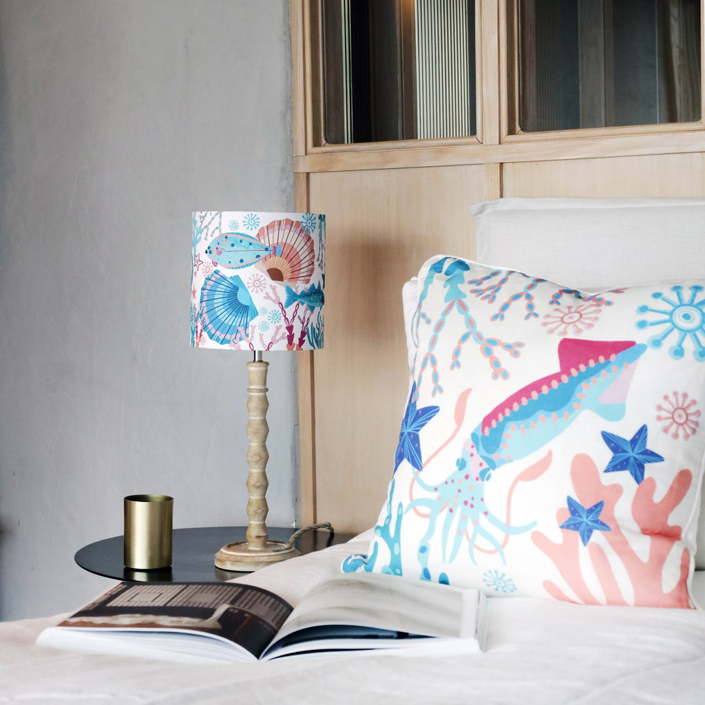 A cozy bedroom setting featuring a marine-themed cushion and matching lamp with colorful illustrations of a squid, fish, starfish, and coral in shades of blue, pink, and orange. The cushion rests on a white bed, while the lamp with a wooden base sits on a black bedside table. A brass candle holder and an open book complete the serene and coastal-inspired decor.