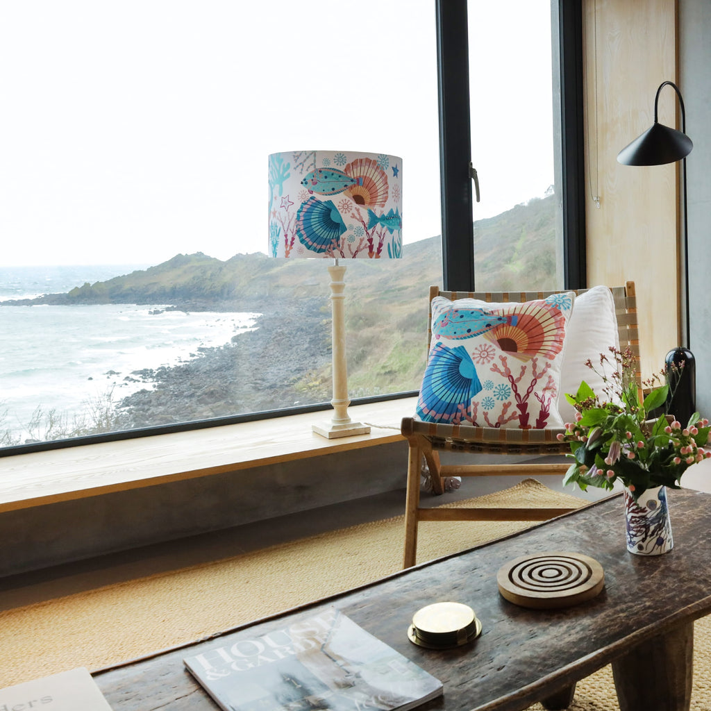 Coastal living space with a large window overlooking the sea. A floor lamp with a marine-themed shade and a matching cushion on a woven chair add a nautical touch. A rustic coffee table holds a vase of flowers, a coaster, and a magazine.