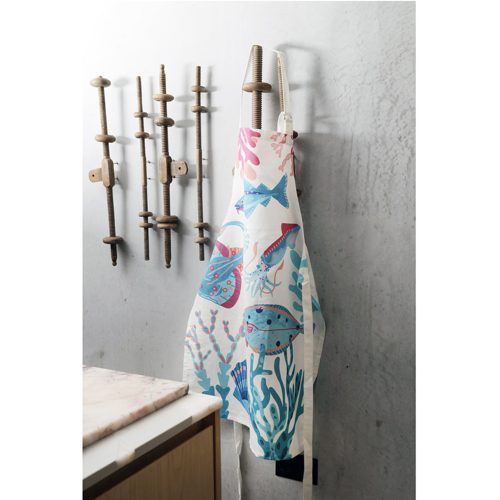A colorful marine-themed apron featuring illustrations of fish, a squid, seaweed, and coral in shades of blue, pink, and turquoise, hanging on a wooden wall-mounted rack with vintage-style wooden clamps. The apron is displayed against a textured concrete wall, with part of a marble countertop visible in the foreground.