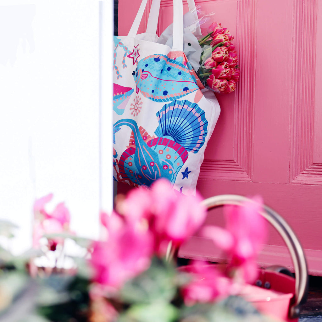 A colorful tote bag with a marine-themed design featuring fish, seashells, and coral in shades of blue, red, and orange, hanging on a pink door. A bouquet of pink tulips peeks out from the bag. In the foreground, blurred pink flowers and a wicker basket handle add depth to the composition.