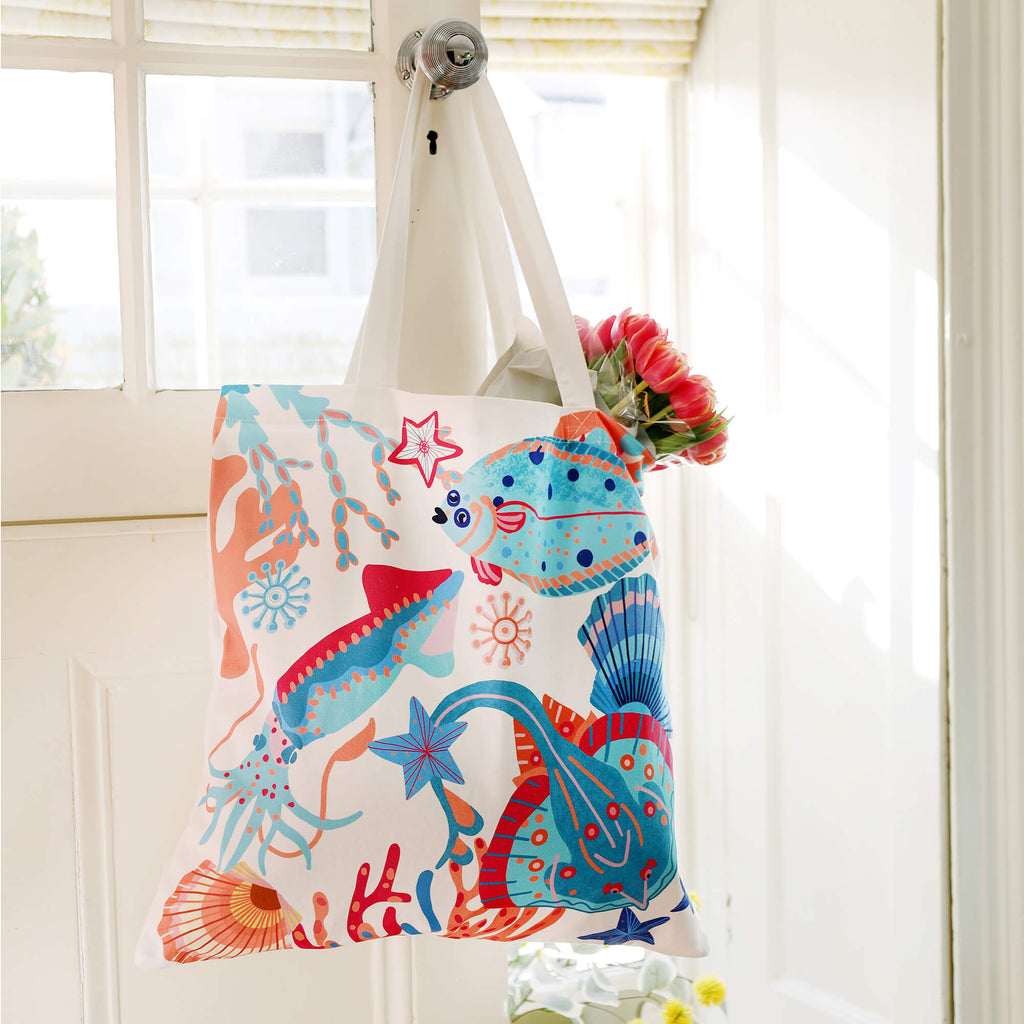 A vibrant tote bag with an ocean-inspired design featuring colorful fish, coral, seaweed, and seashells in shades of blue, red, and orange. The bag hangs on a white door handle, with a bouquet of pink tulips peeking out from the top. Sunlight streams through a window in the background, casting a bright and airy feel.