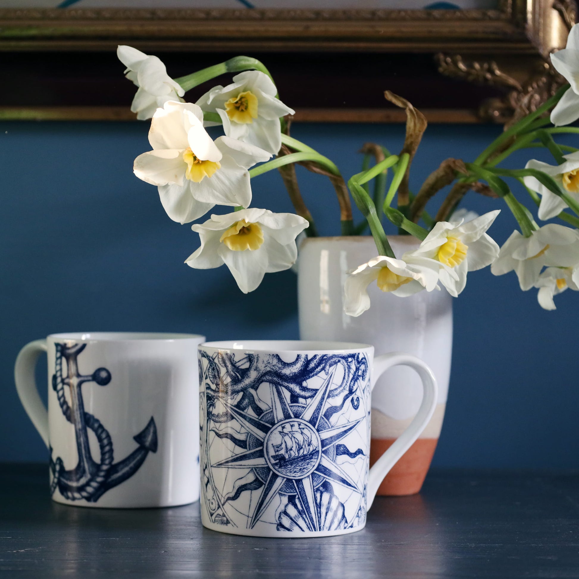 Two mugs with decorative designs and a vase of white flowers on a dark surface.