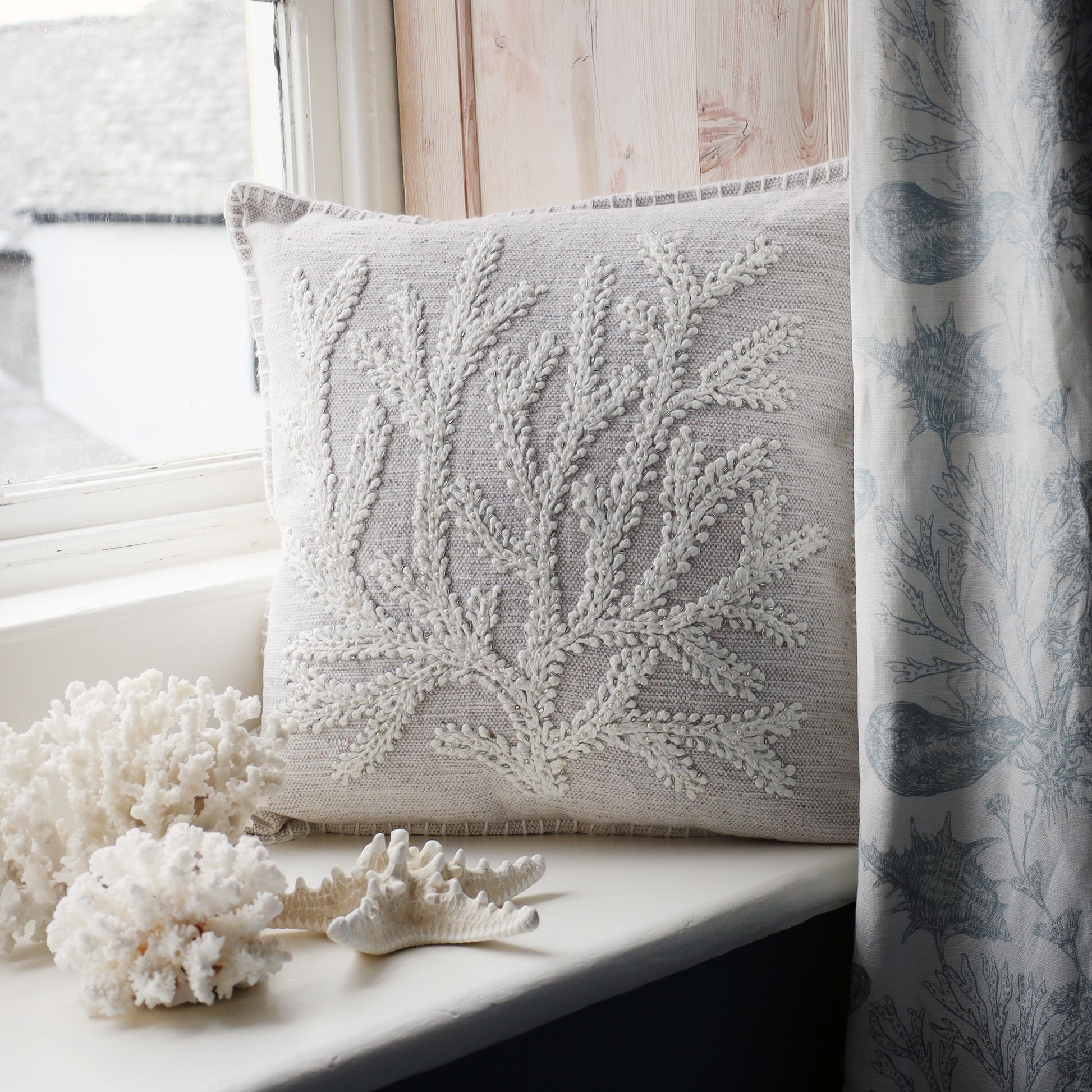 Decorative pillow with coral pattern on a windowsill next to decorative corals and a curtain.