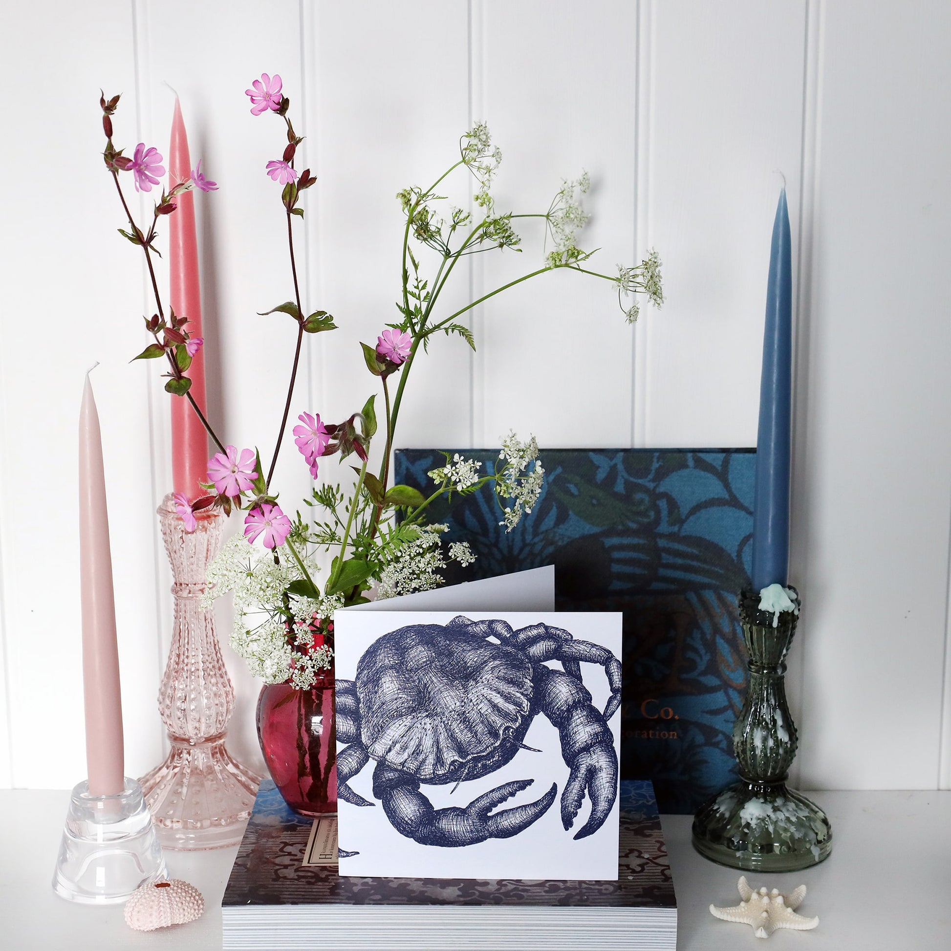 greeting card with navy illustrated crab on a white background on shelf with pink and blue candles in candlesticks and a small cranberry glass jug with wild flowers in