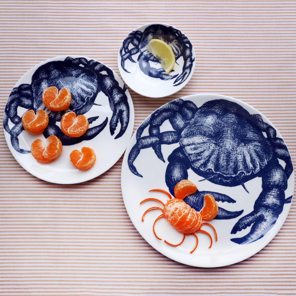 Three different sized white bone china plates with an illustrated navy blue crab on them. The largest plate has a crab made from a satsuma on it and the other 2 have a crab claws made from a satsuma and a slice of lemon on it. Both plates are shot from above on a striped yellow gold cloth.