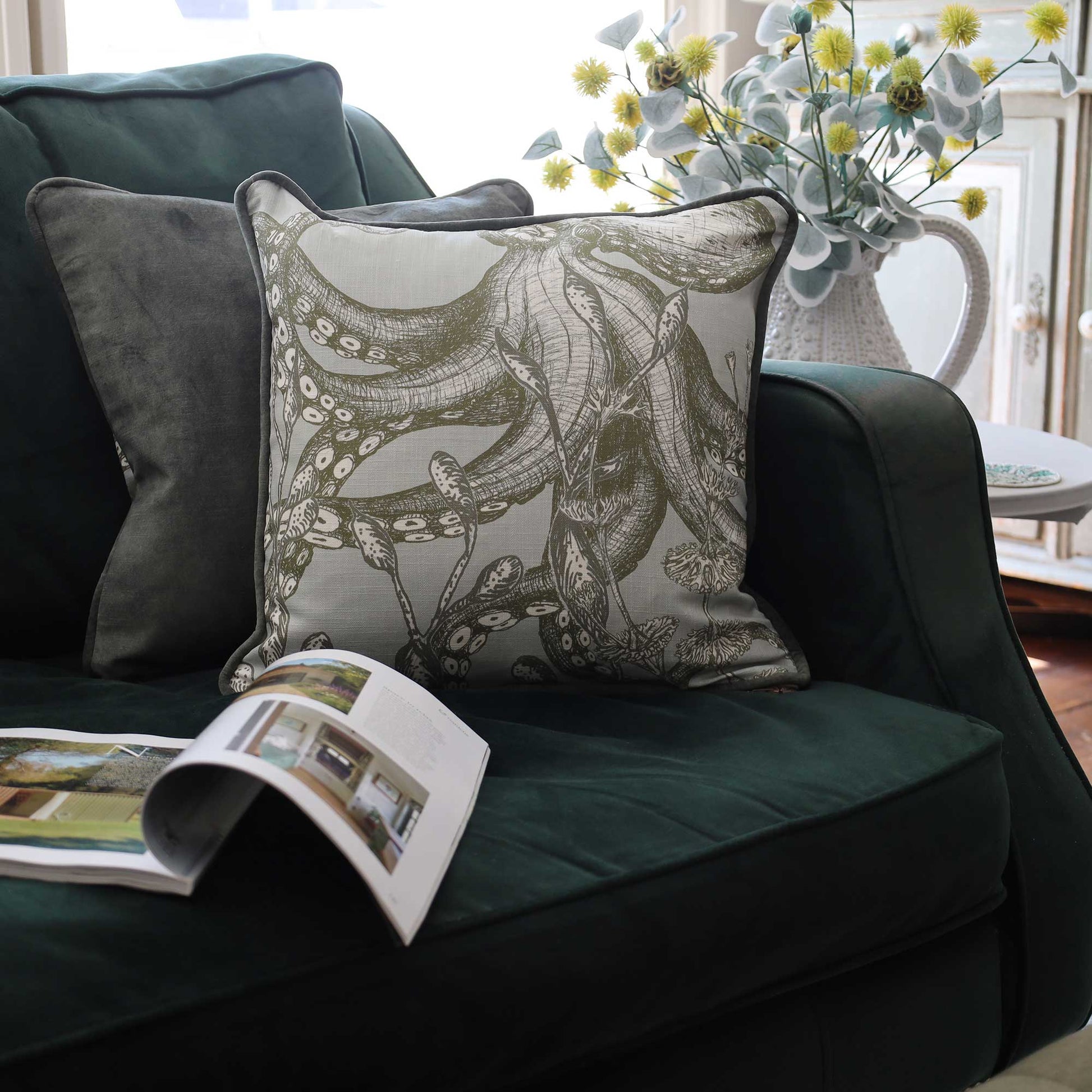 A square decorative throw pillow with an octopus tentacle pattern in muted gray and olive tones, styled on a dark green velvet sofa next to a dark gray pillow, with an open magazine and a vase of yellow flowers in the background.
