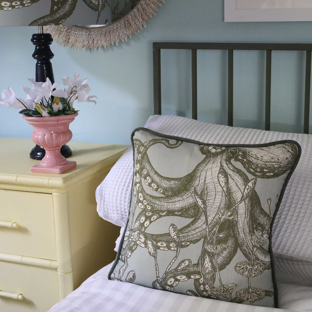 A square decorative throw pillow featuring a detailed octopus illustration in olive green on a light beige background, styled on a bed next to a pale yellow nightstand with a pink vase of flowers.