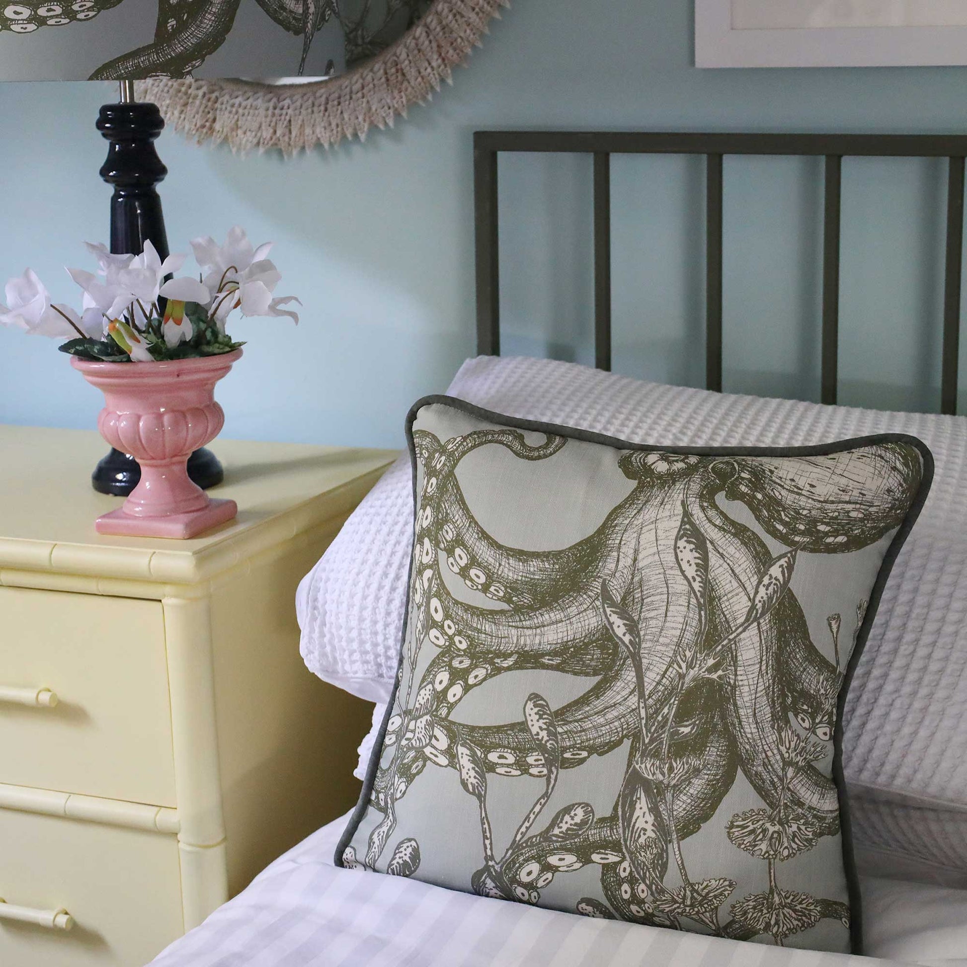 A square decorative throw pillow featuring a detailed octopus illustration in olive green on a light beige background, styled on a bed next to a pale yellow nightstand with a pink vase of flowers.