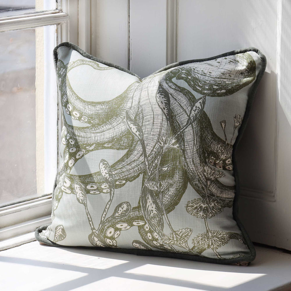A square decorative throw pillow with a detailed octopus and botanical illustration pattern in olive green and gray on a light cream background, styled on a windowsill.