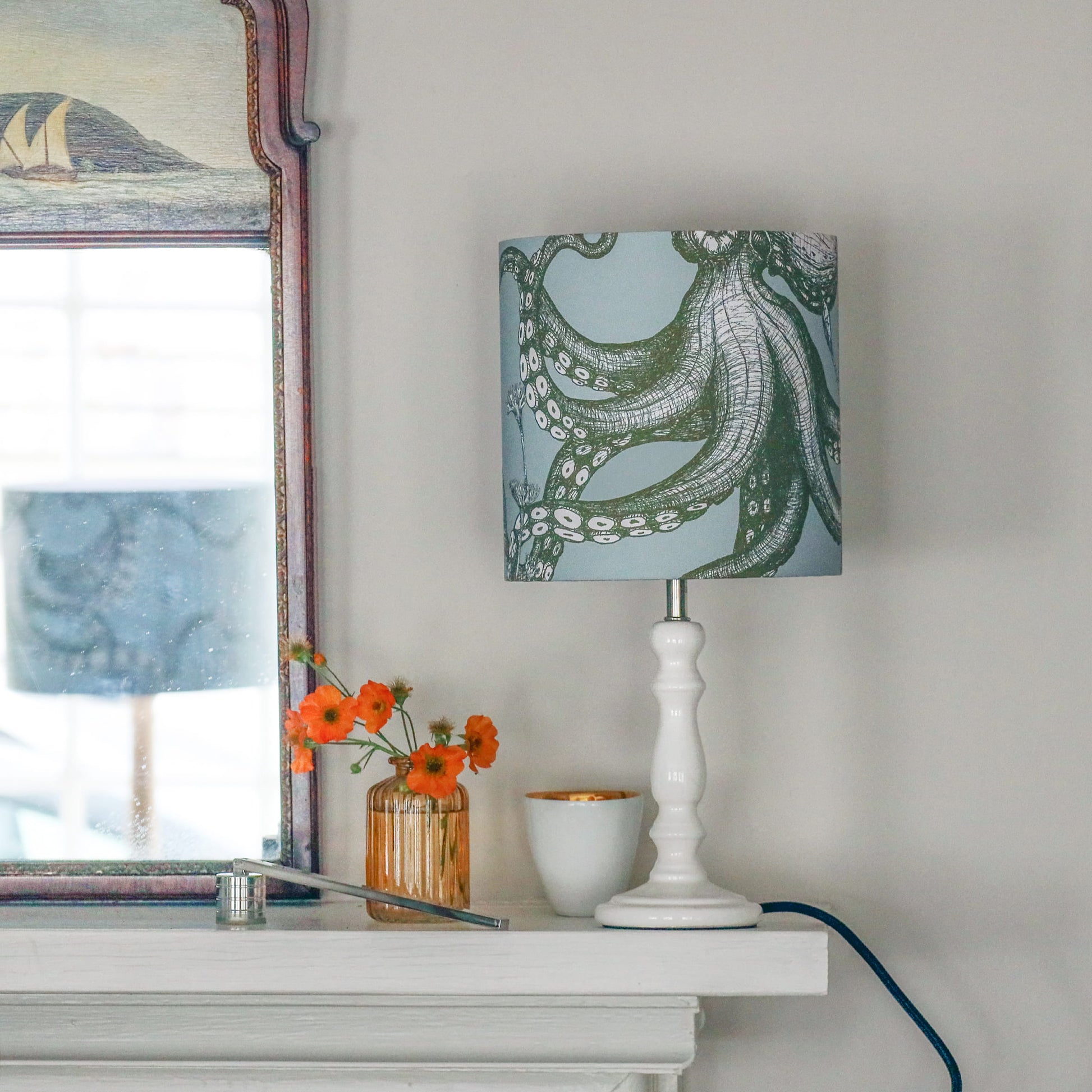 Lamp with a decorative shade on a white surface next to a mirror and flowers.
