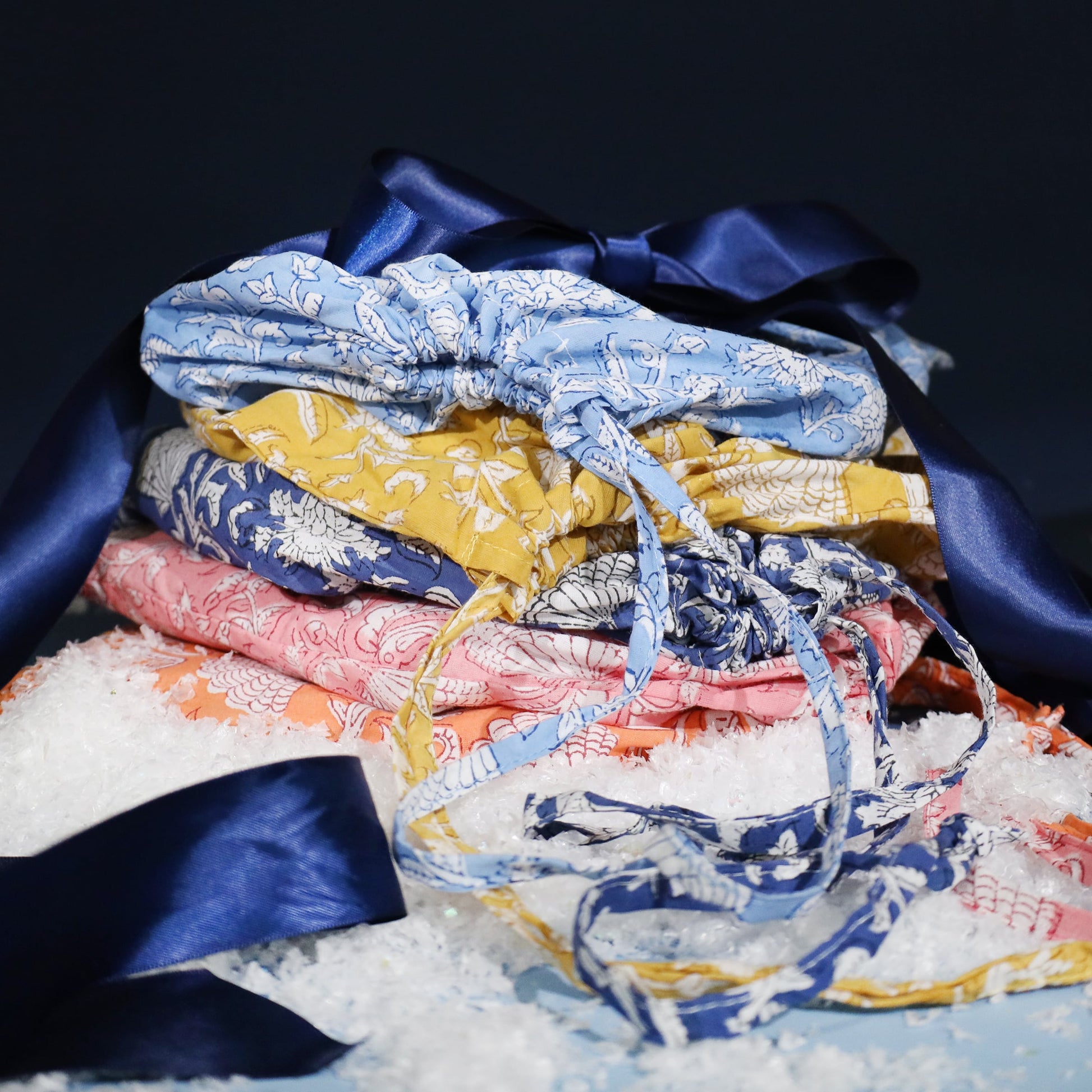Stack of colourful block-printed cotton dressing gowns in blue, mustard, navy, pink, and coral tones, tied with a navy satin ribbon and surrounded by faux snow for a festive presentation.
