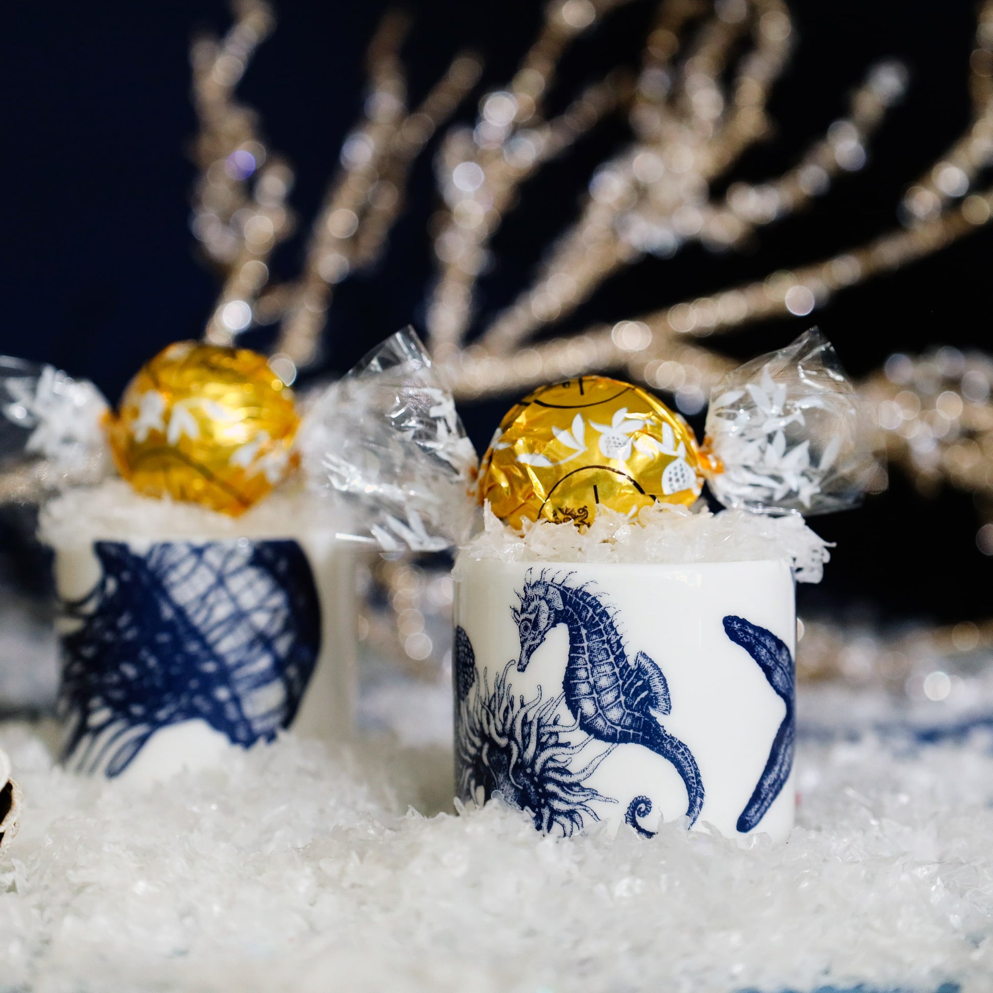 Festive scene with two white egg cups decorated with blue marine illustrations of a seahorse and jellyfish, each holding a gold-wrapped chocolate truffle nestled in faux snow, with sparkling golden branches softly blurred in the background.