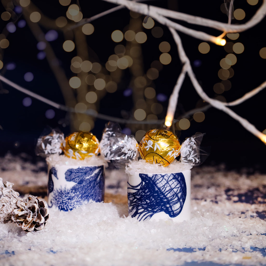 Two white egg cups decorated with blue sea creature illustrations, each holding a gold-wrapped chocolate truffle nestled in faux snow, surrounded by pinecones and twinkling fairy lights on white branches against a dark festive background.