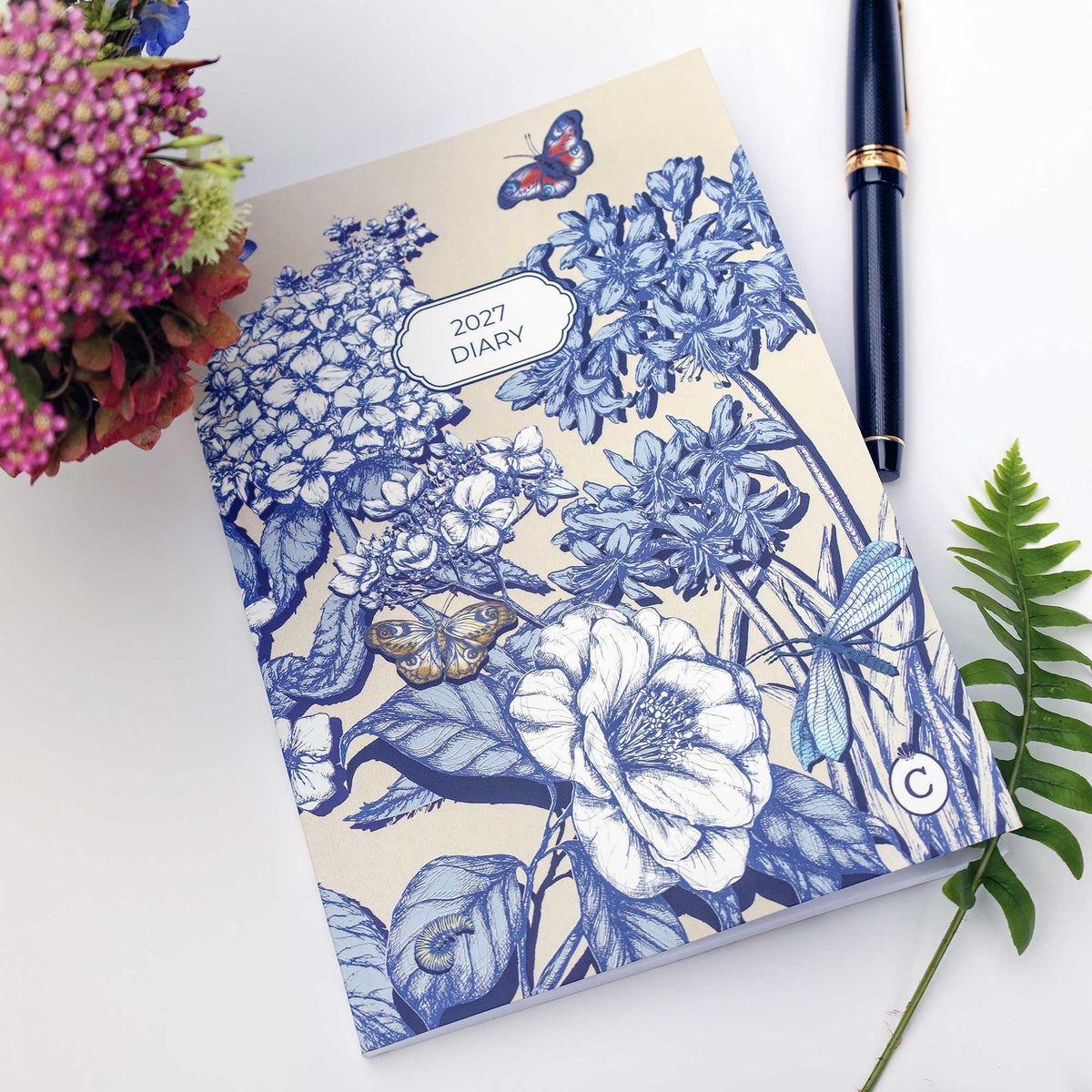 A 2027 floral diary with a blue and cream botanical illustration, styled on a white surface with a black pen, flowers, and a green fern.