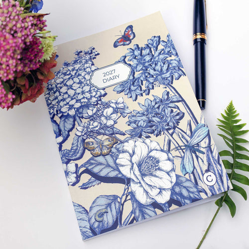 A 2027 floral diary with a blue and cream botanical illustration, styled on a white surface with a black pen, flowers, and a green fern.