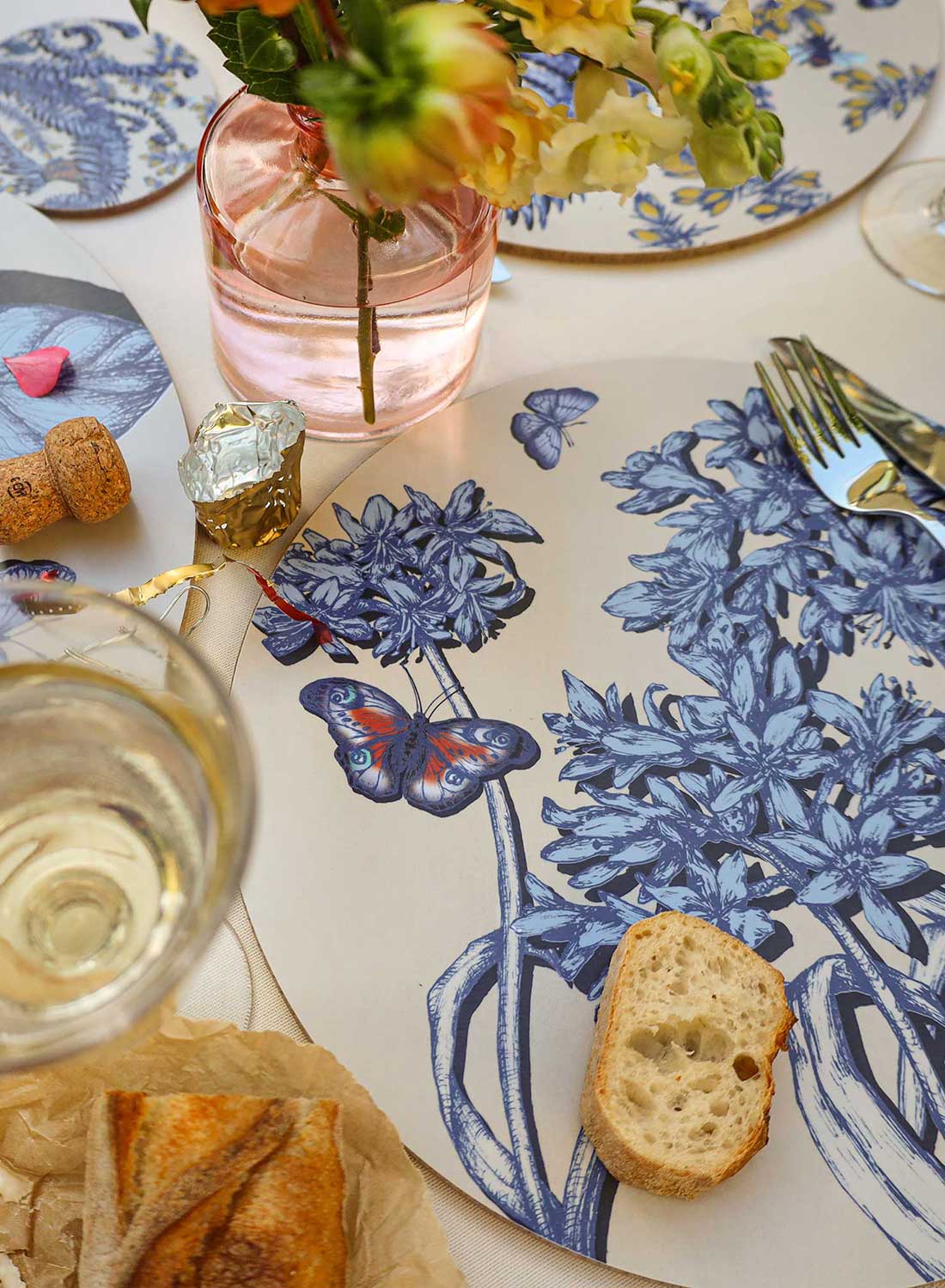 Overhead view of an elegant table setting featuring round placemats with intricate blue botanical illustrations of flowers, leaves, ferns, butterflies and insects on a cream background. A vibrant floral centrepiece of orange dahlias, delicate striped cream flowers and bright orange blooms adds colour to the arrangement. Bamboo-handled cutlery, matching botanical coasters, glasses of white wine, a bowl of green olives, crusty bread and soft cheese drizzled with olive oil complete the setting on a light linen