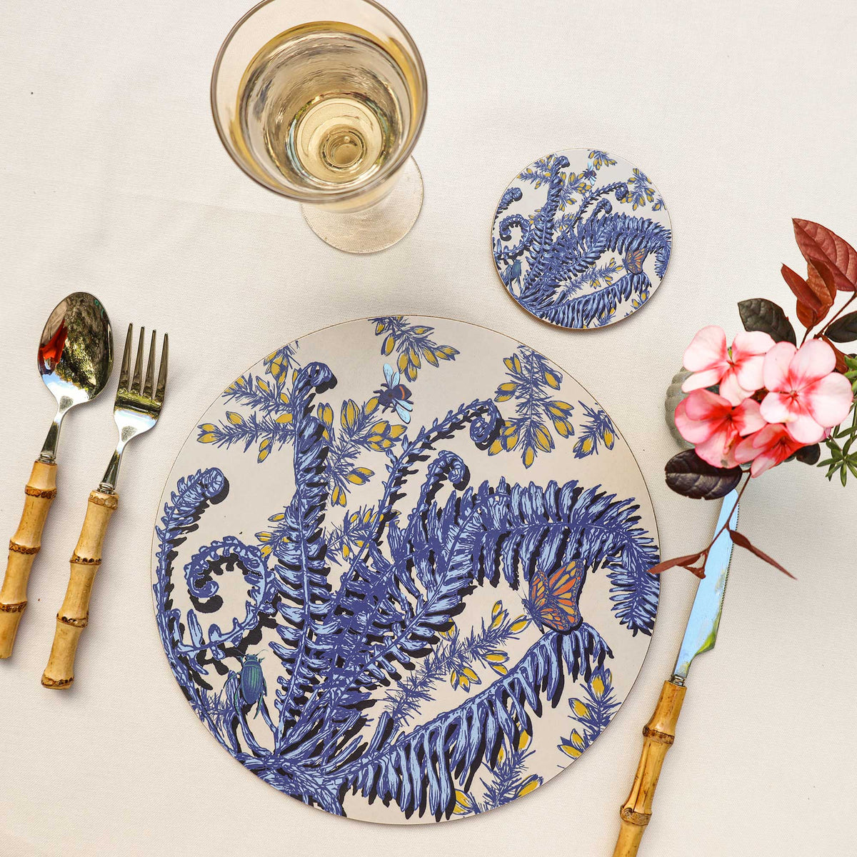 
Overhead view of a round cream placemat featuring intricate blue botanical illustrations of ferns, curled fronds and yellow flowers, accented with a colourful orange butterfly, blue bee and green beetle. Styled on a neutral linen tablecloth with a matching botanical coaster, a glass of white wine, bamboo-handled cutlery and a small arrangement of pink flowers with burgundy leaves.