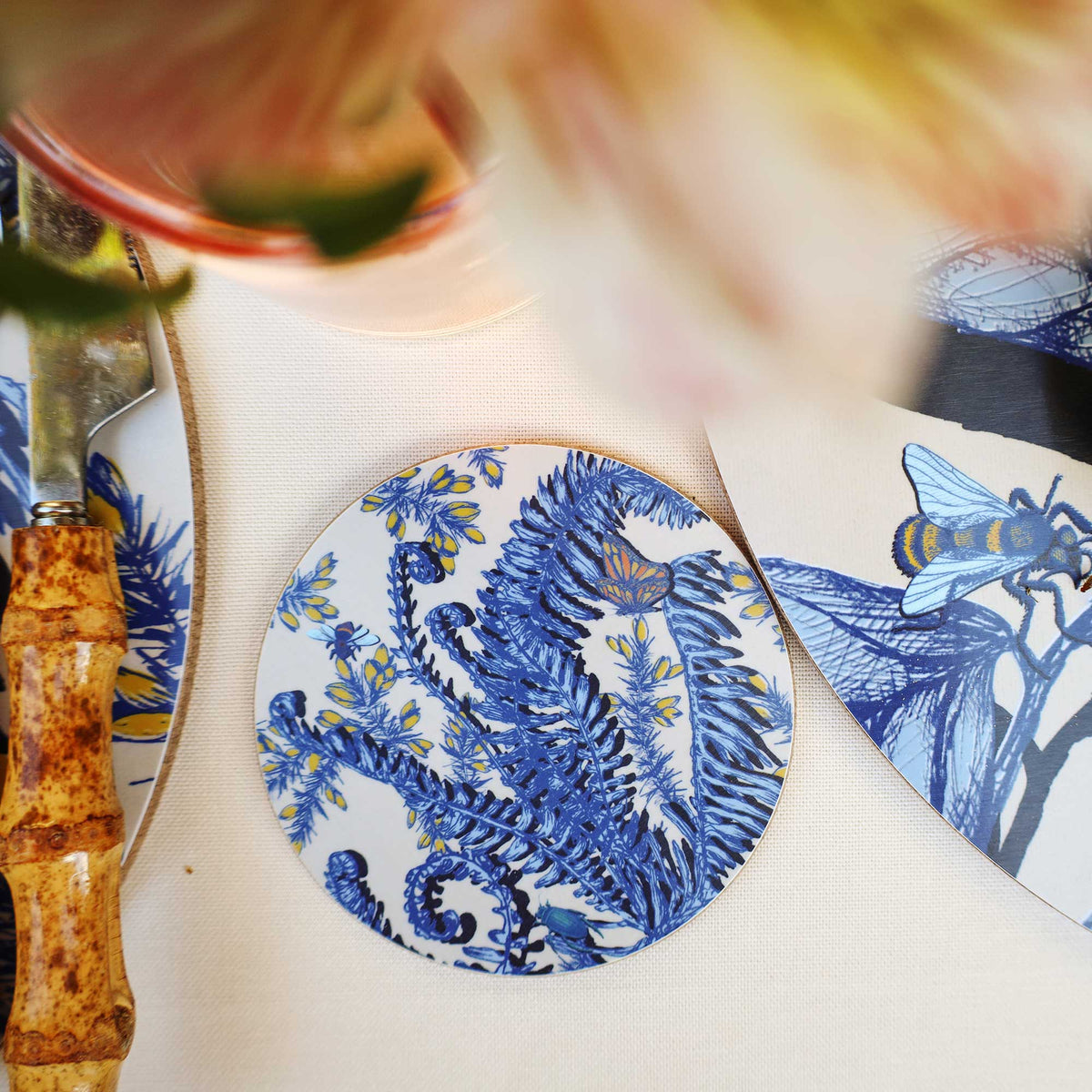 Cream Cornwall round coaster featuring intricate blue fern illustrations, delicate yellow flowers, and colourful butterflies and insects on a cream background, styled on a neutral linen tablecloth alongside bamboo-handled cutlery, a pink drink and matching botanical tableware.