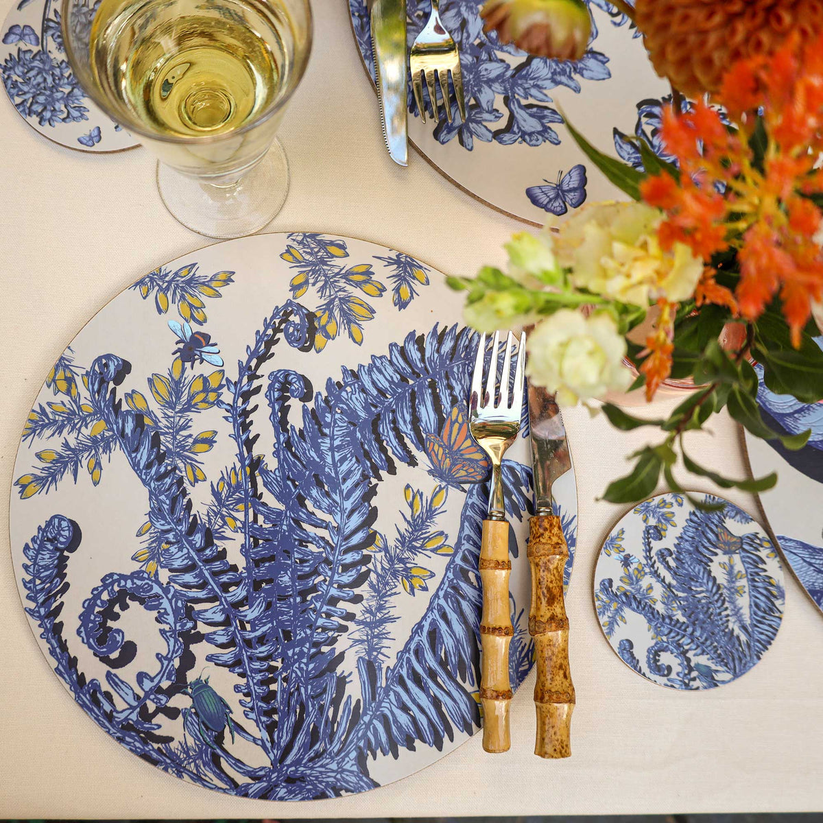 Elegant table setting featuring a Cream Cornwall round placemat and matching coaster, decorated with intricate blue fern illustrations, yellow flowers, butterflies and insects on a cream background. Styled with bamboo-handled cutlery, a glass of white wine, coordinating botanical tableware and a vibrant bouquet of orange and cream flowers.