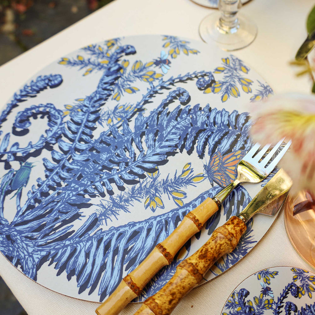 Close-up of a round cream placemat featuring intricate blue botanical illustrations of ferns, curled fronds and delicate yellow flowers, accented with an orange and blue butterfly, a blue bee and a green beetle. Styled on a neutral linen tablecloth with bamboo-handled cutlery, a matching botanical coaster and a wine glass, with softly blurred flowers in the foreground.