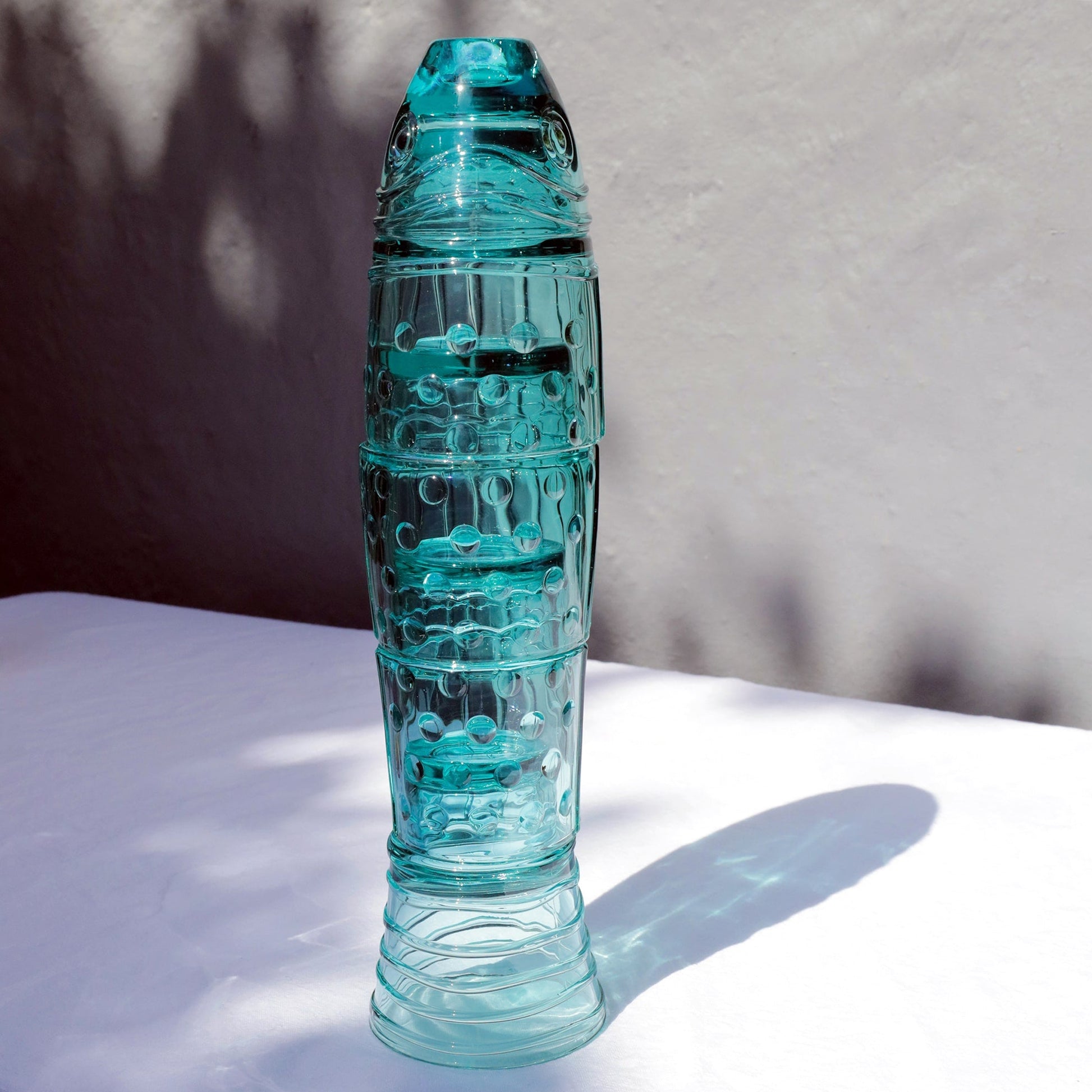Turquoise stacking fish glasses forming the shape of a fish, displayed on a white surface with sunlight and shadow.