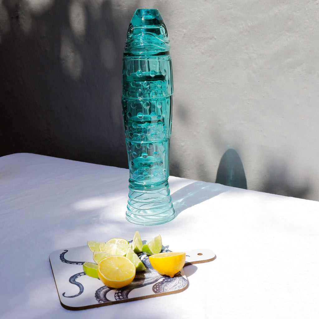 A turquoise glass stacking fish beaker set is arranged on a white tablecloth beside a small cutting board with an octopus design, holding sliced lemons and limes. Sunlight creates soft shadows on the wall and table.