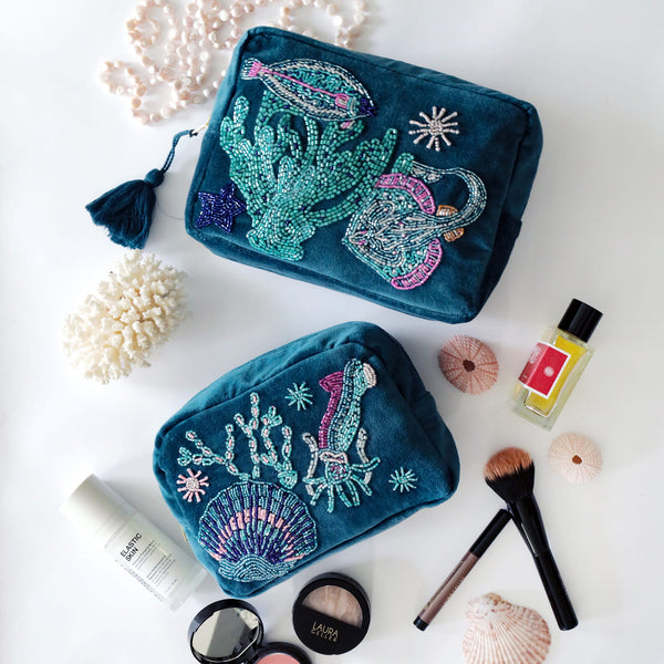 Two teal beaded pouches with sea-themed designs on a white surface with makeup items.
